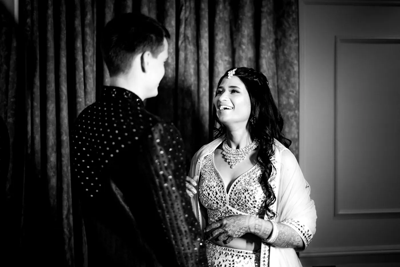 In black and white, Medha laughs up at Stefan, who looks down at her, against dark curtains