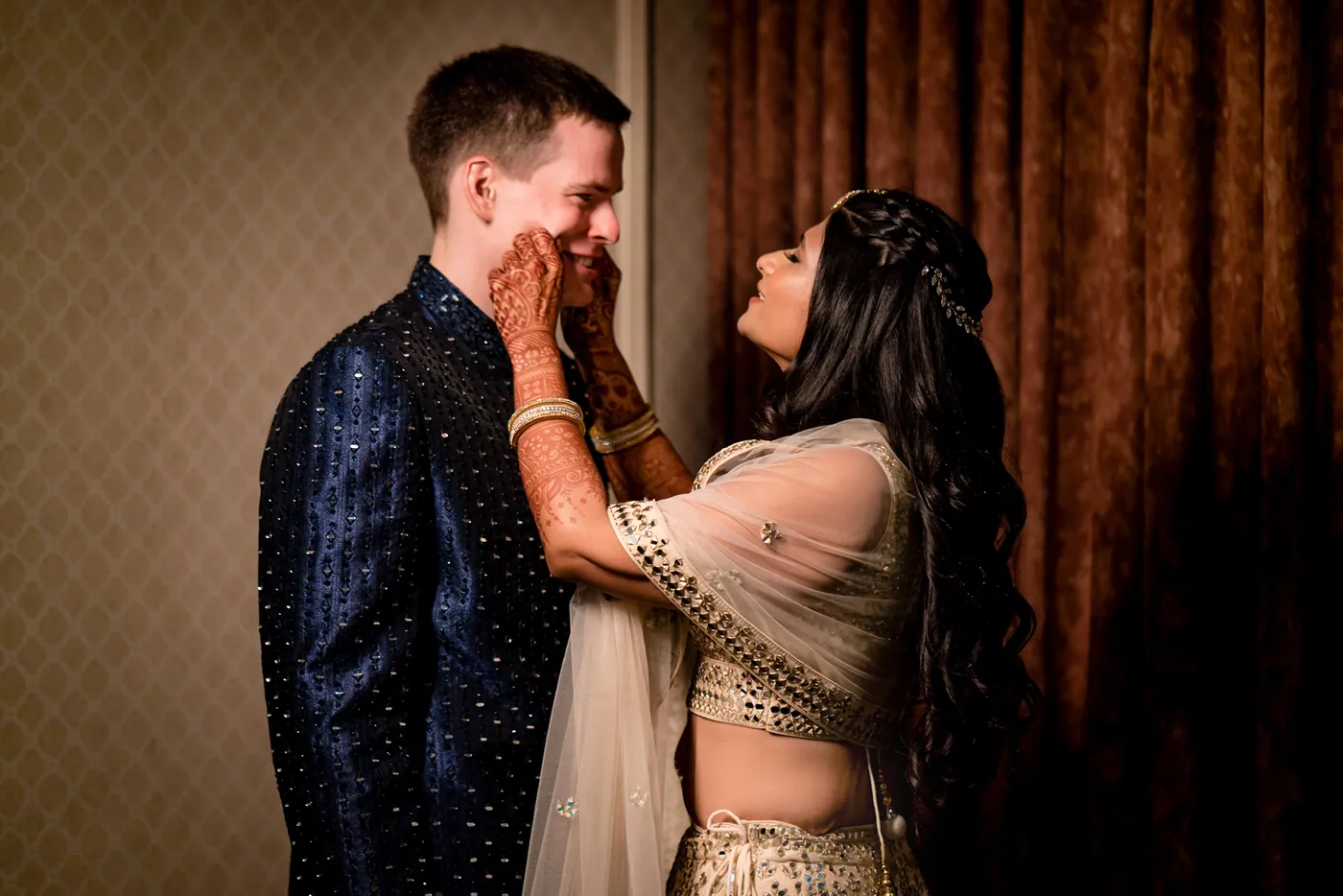 Medha reaches up to hold Stefan's face and laughs while he smiles, a brown curtain behind them