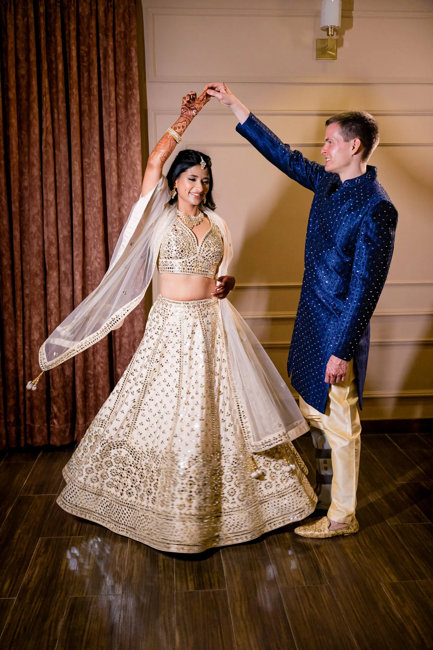 Stefan raises a hand as Medha twirls in her cream lehenga, her dupatta flaring across the wooden floor