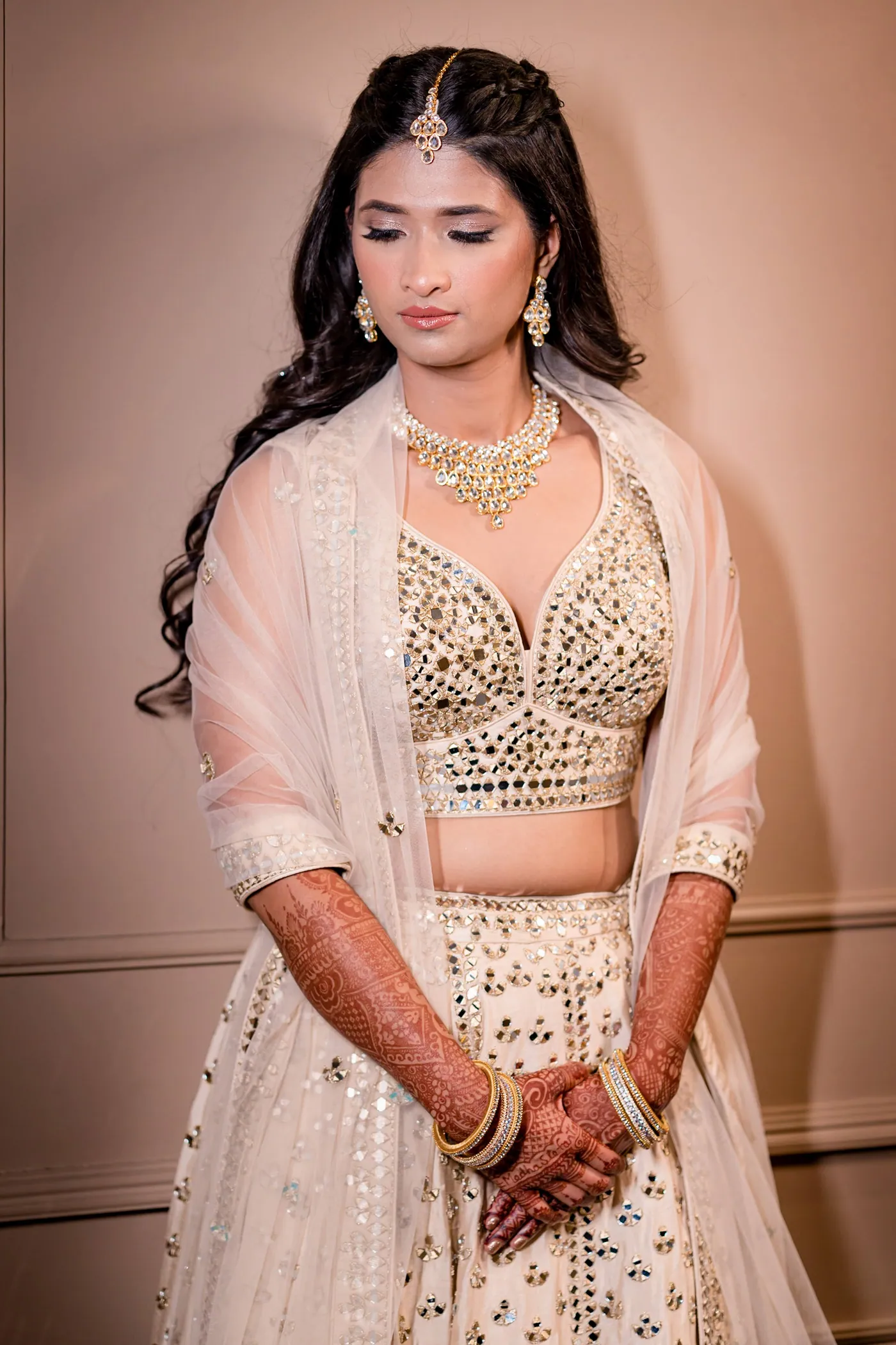 Medha looks down with a quiet smile, her hennaed hands folded in front of her cream lehenga