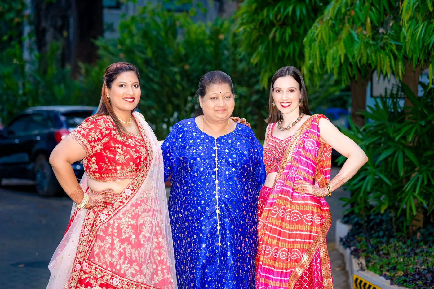 Three women in red, blue and pink outfits pose with hands on hips in front of a hedge