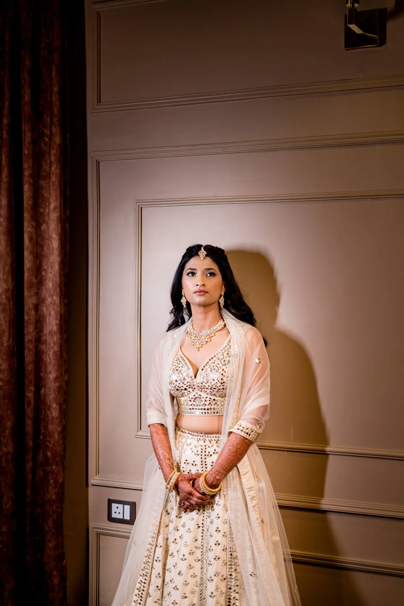 Medha stands alone against a cream panelled wall in her lehenga, her shadow falling beside her