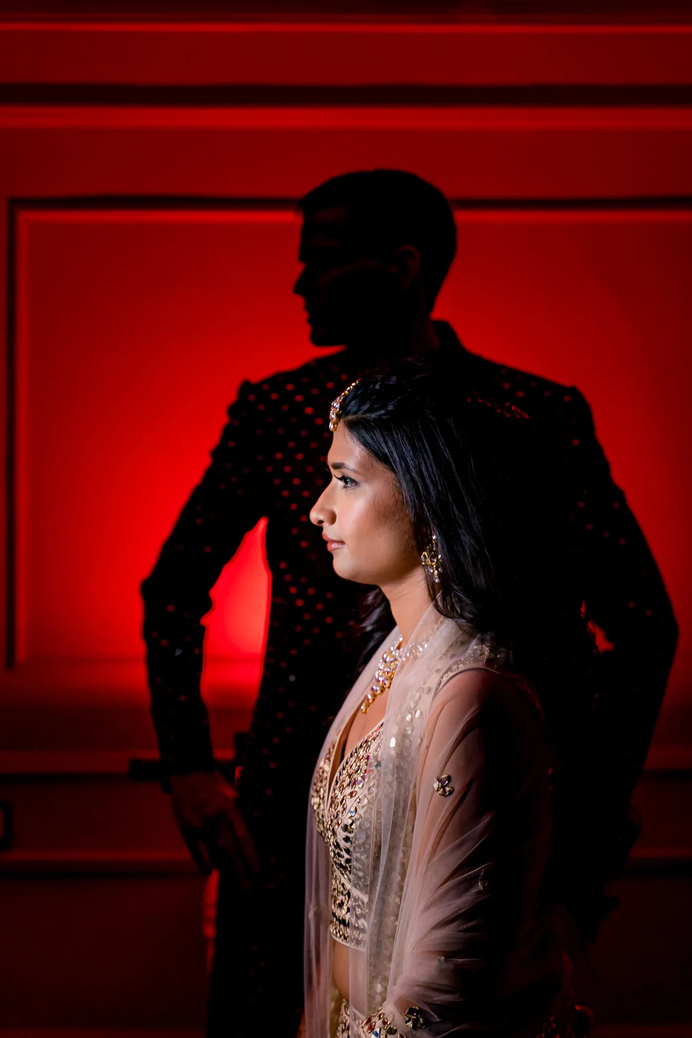 Against a deep red wall, Stefan stands as a dark shape behind Medha, who gazes aside