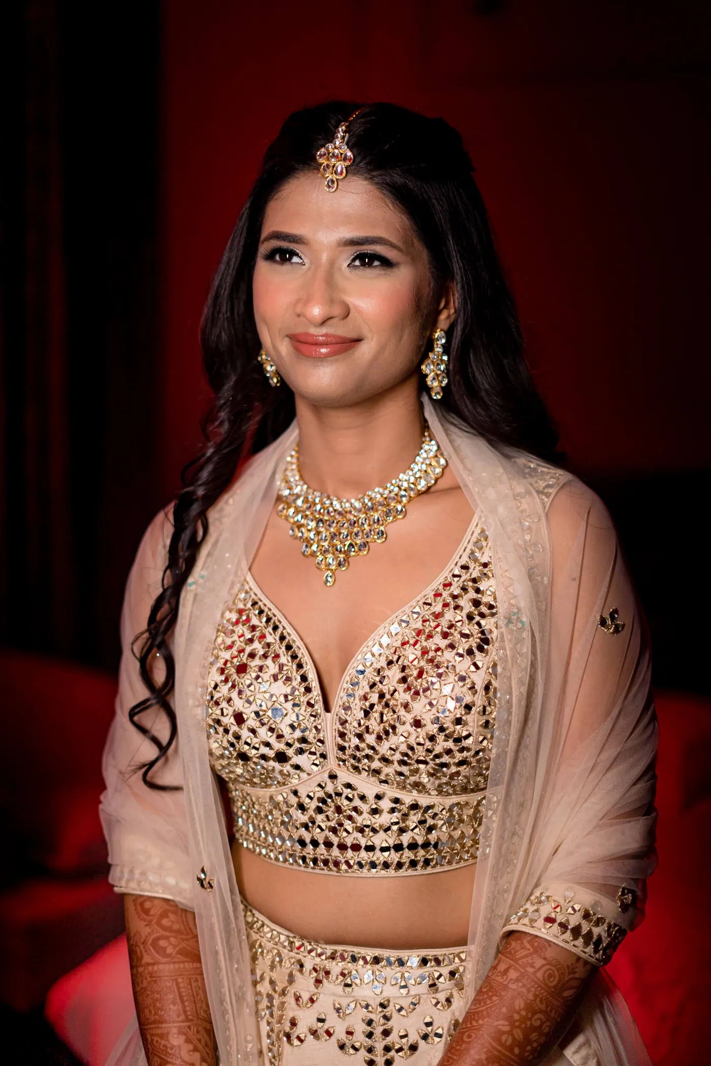 Medha gazes aside with a soft smile in a gold tikka and a crystal necklace, red glow behind