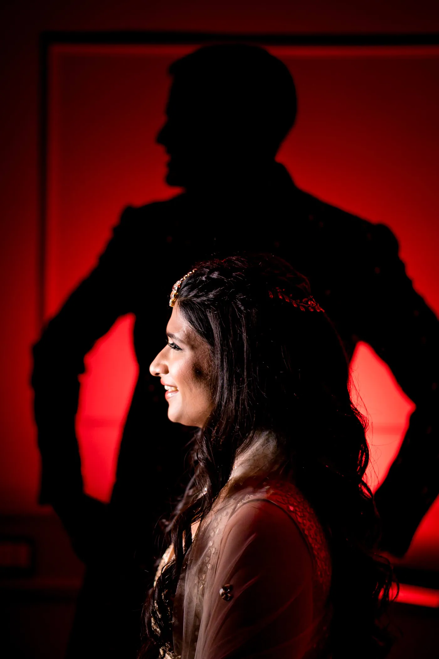 Medha looks to one side with a bright smile while Stefan stands behind her as a dark outline against red