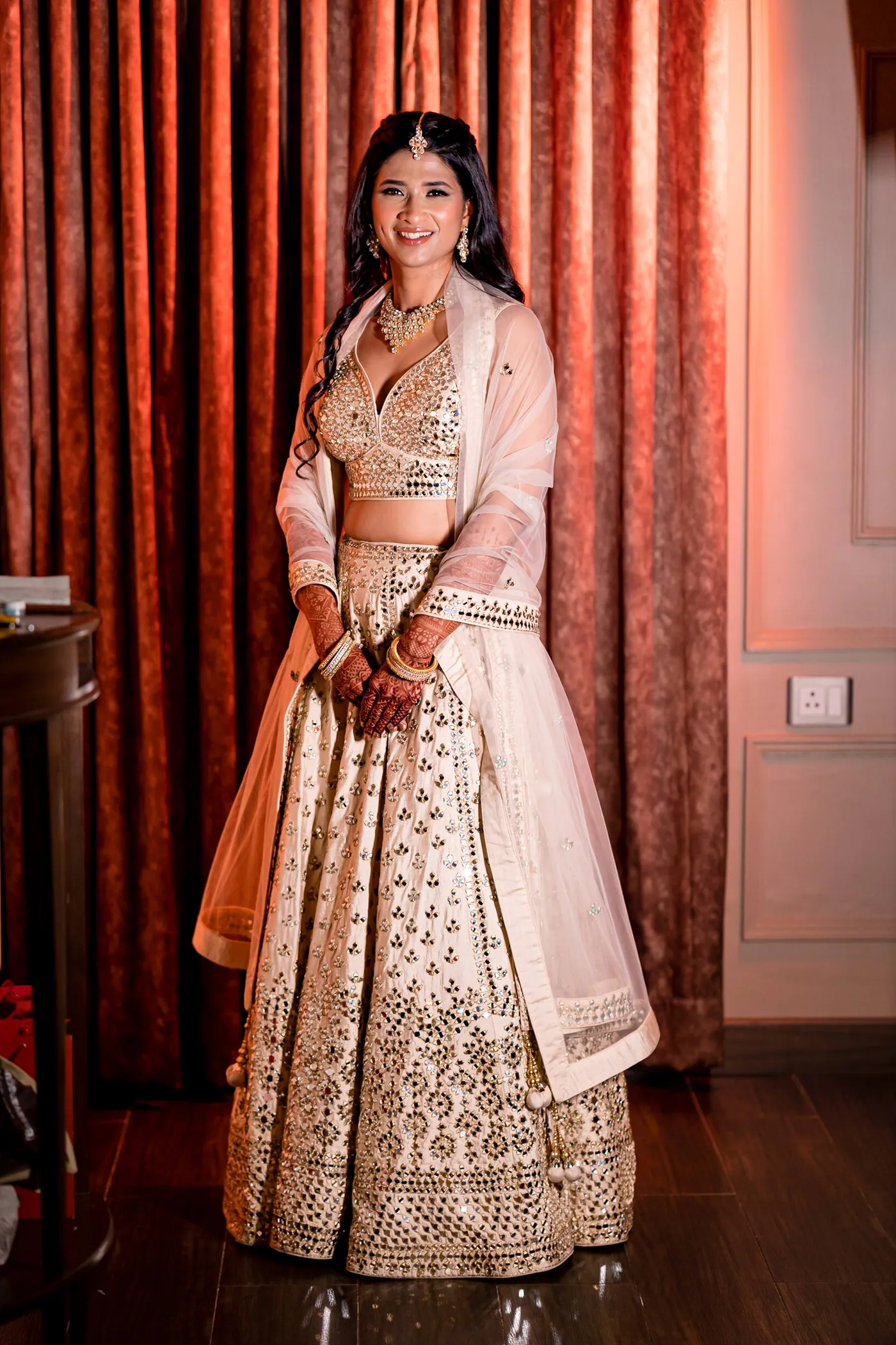 Medha stands full length in her cream and gold lehenga, orange curtains falling behind her