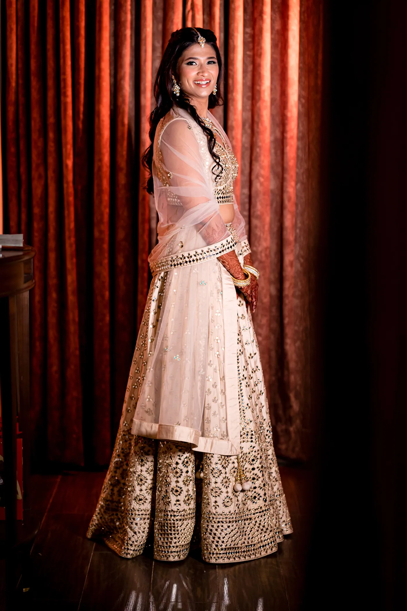 Medha glances back over her shoulder with a smile in front of rust coloured curtains
