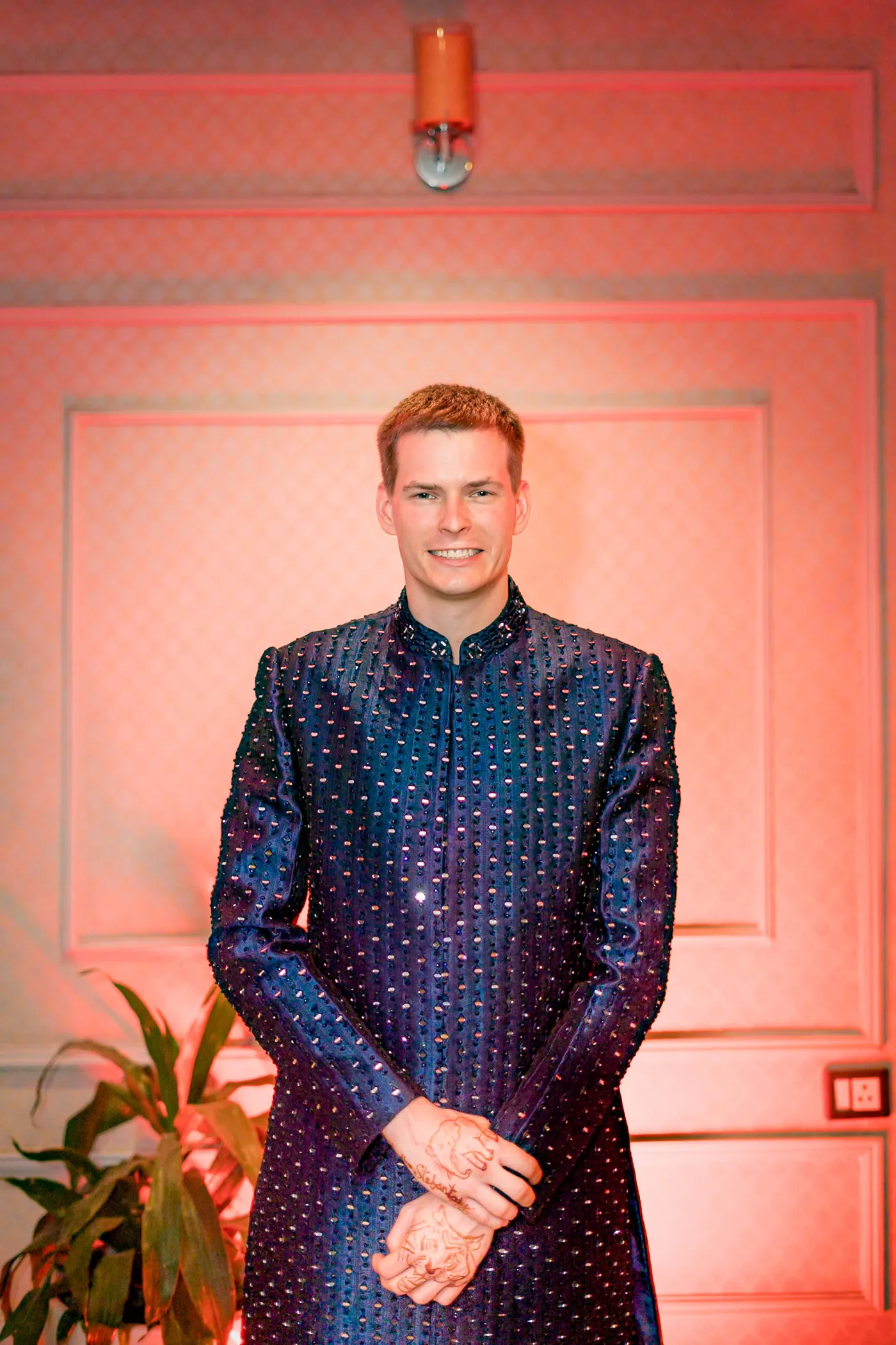 Stefan stands with hands clasped in a navy sherwani against a glowing peach wall