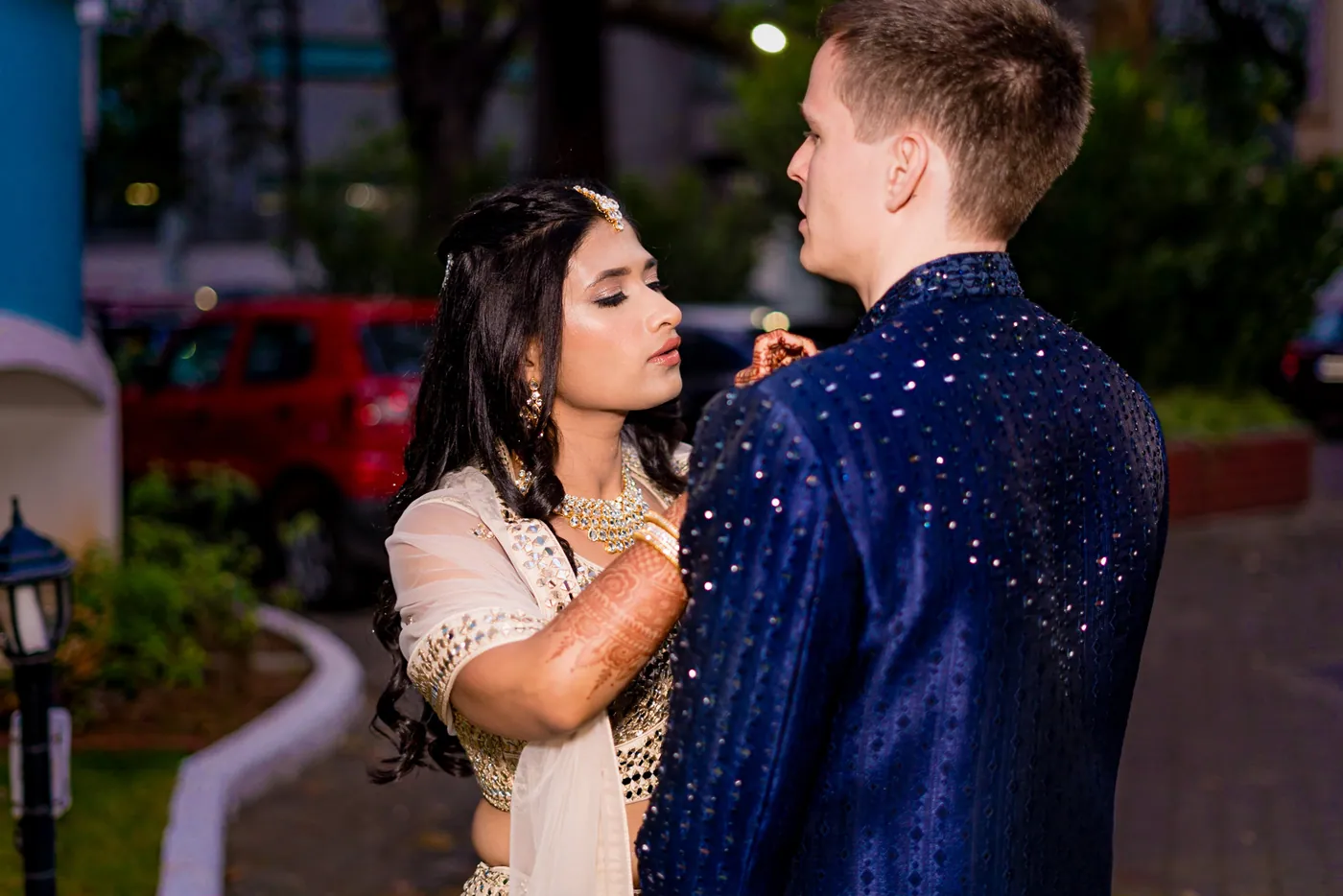 Medha adjusts the lapel of Stefan's sherwani on a quiet path beside the club