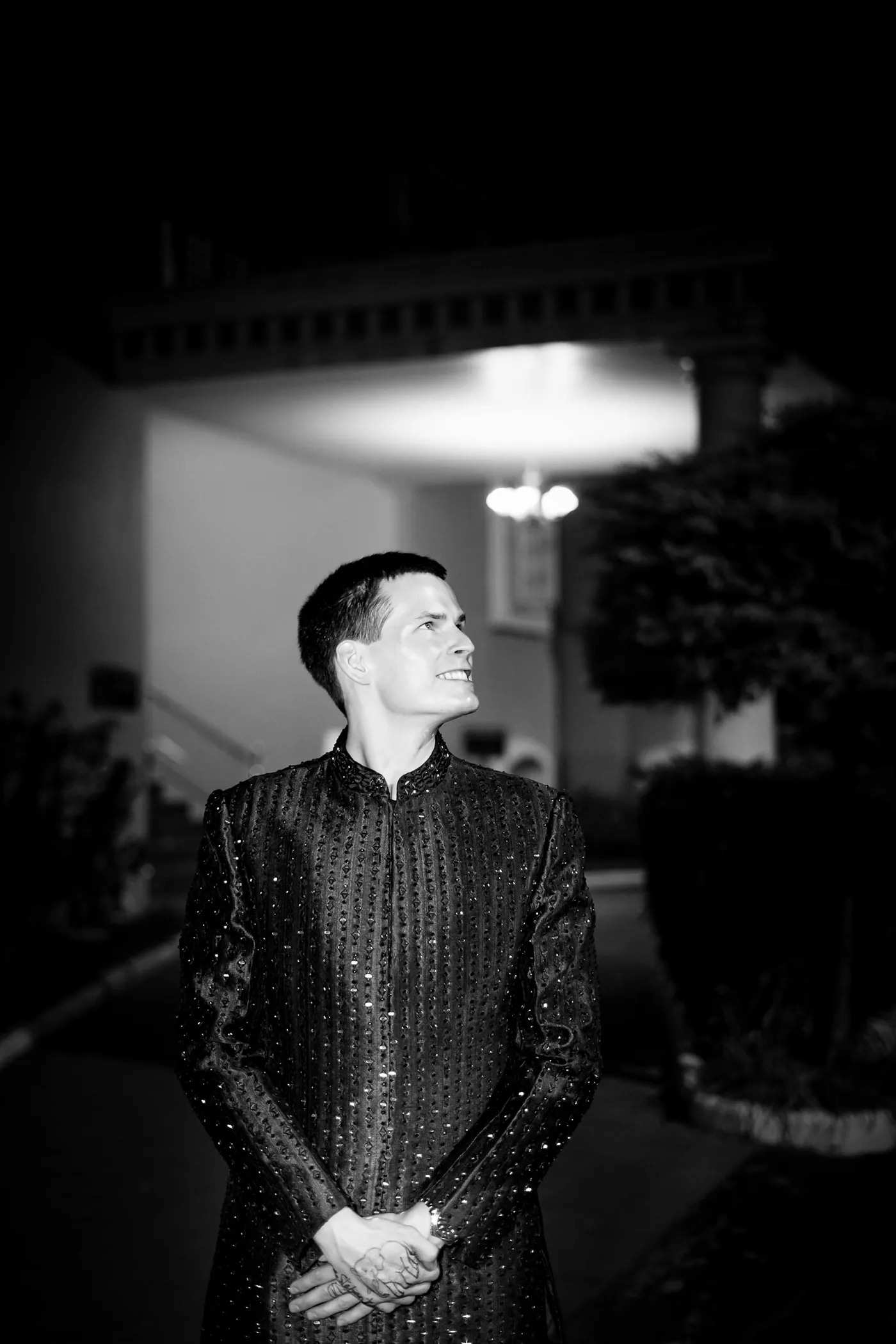 In black and white, Stefan stands with hands clasped on a dark path, looking up and away