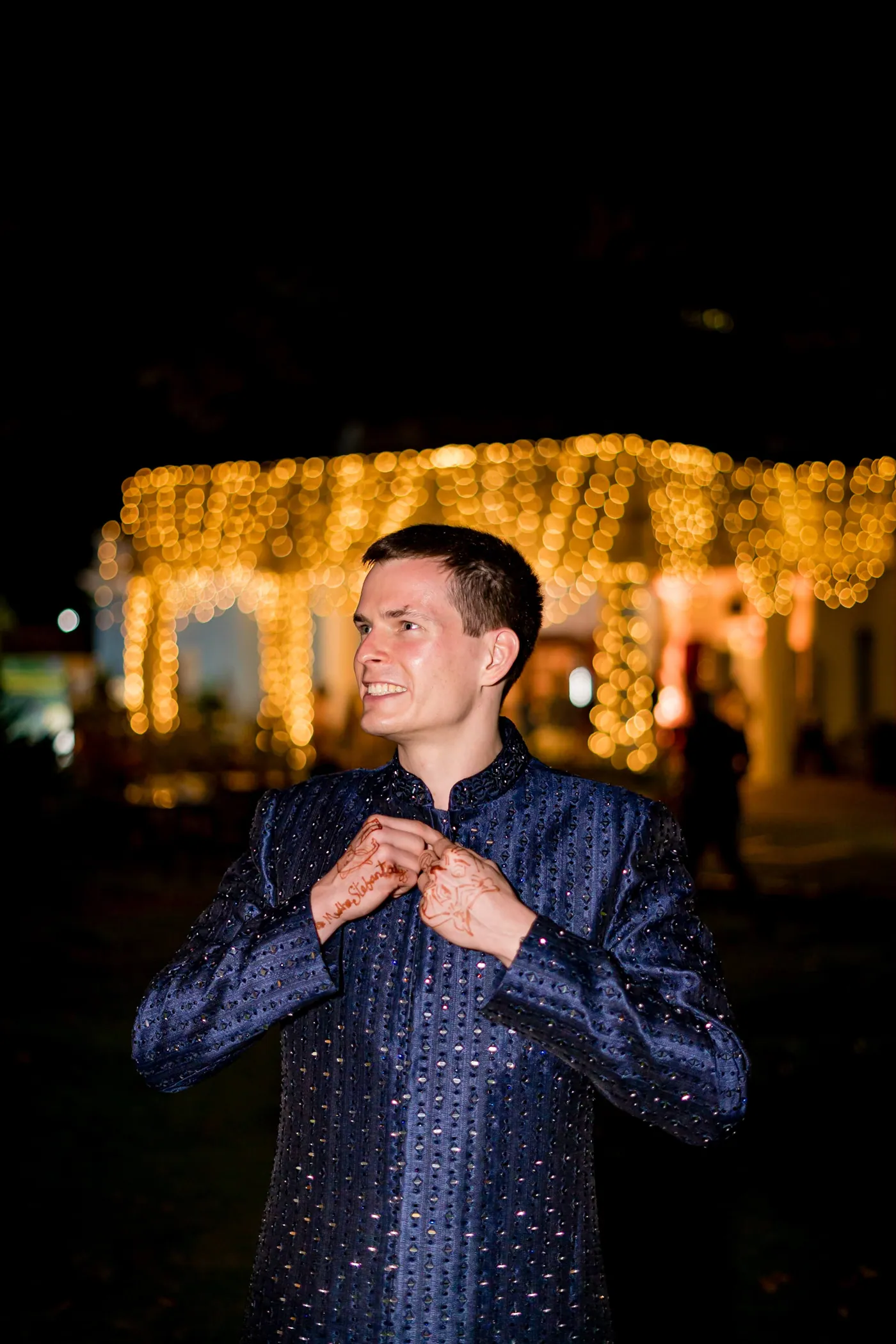 Stefan smiles as he adjusts his navy sherwani cuffs, a canopy of lights glowing behind him