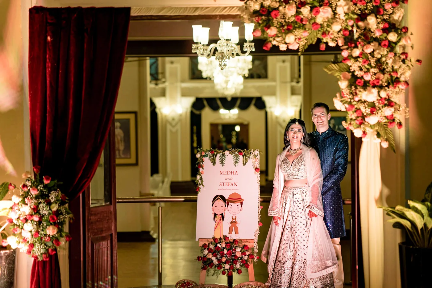 Stefan and Medha stand beside an illustrated welcome sign under a flower arch at a doorway