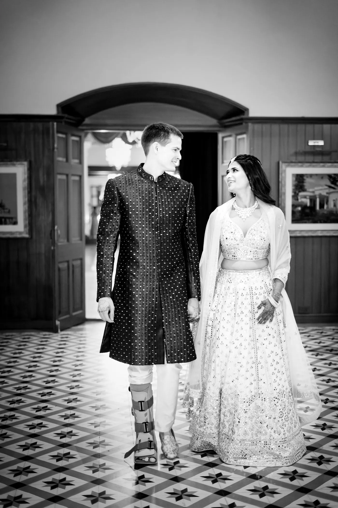 In black and white, Stefan and Medha walk across a tiled floor, his medical boot showing