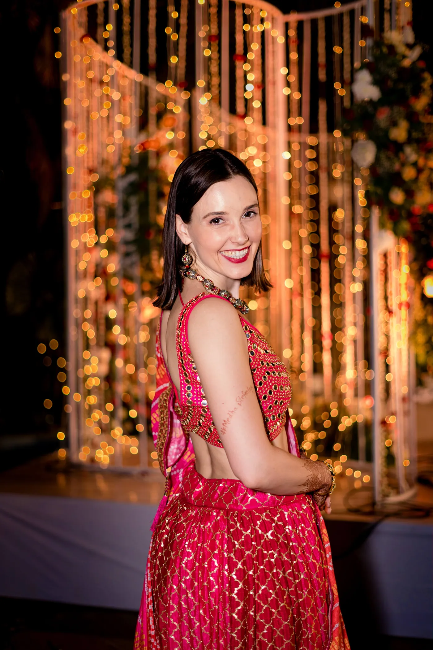 Stefan's sister turns and smiles over her shoulder in a red lehenga against warm glowing lights