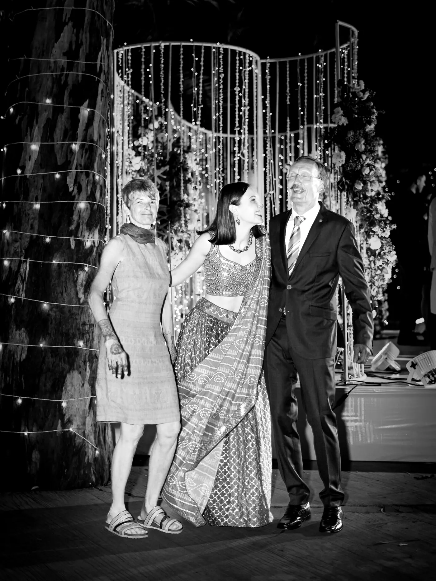 Stefan's parents stand either side of a smiling woman in a patterned lehenga in front of the light curtain
