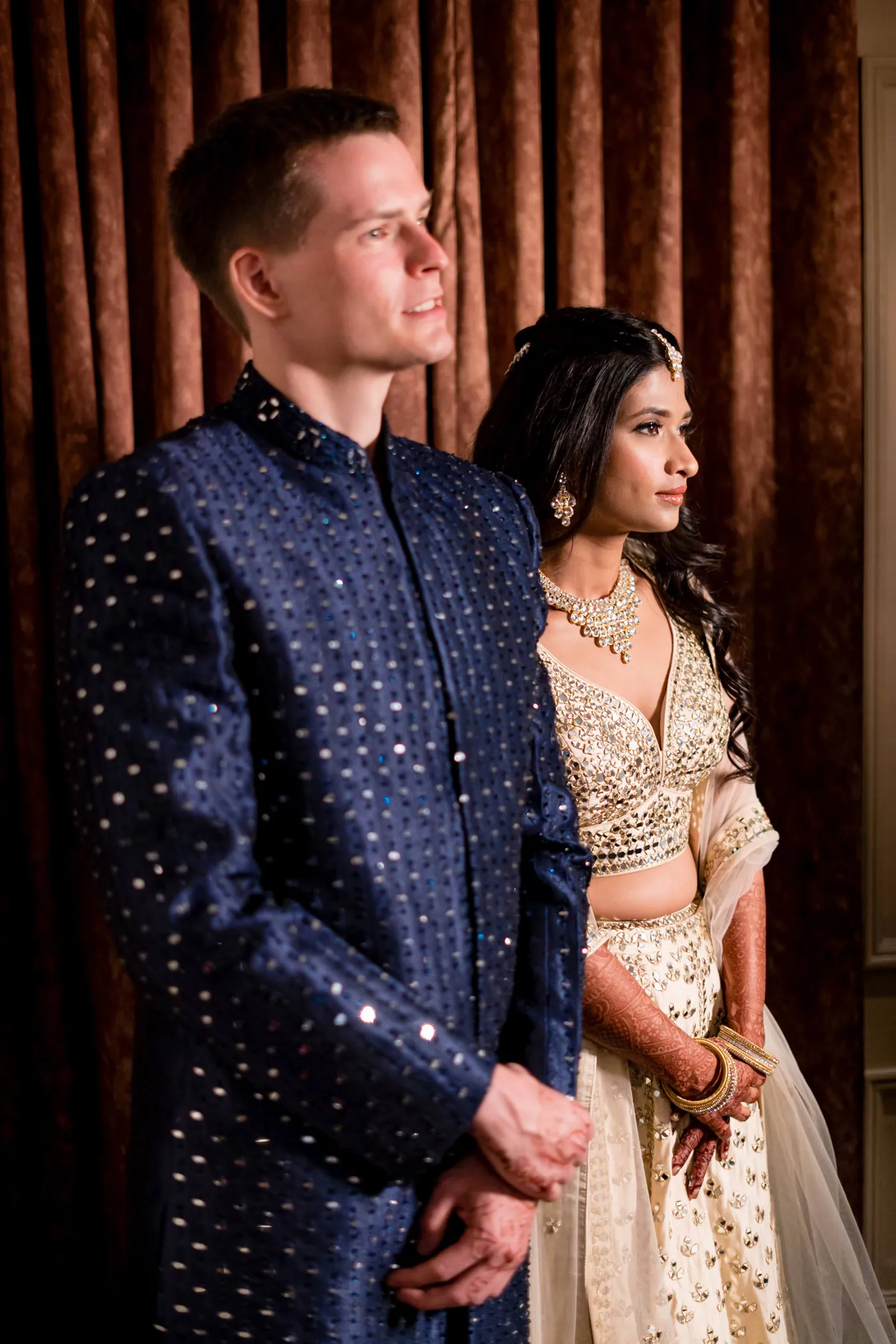 Stefan looks off to the side in his navy sherwani as Medha stands close behind him against dark drapes