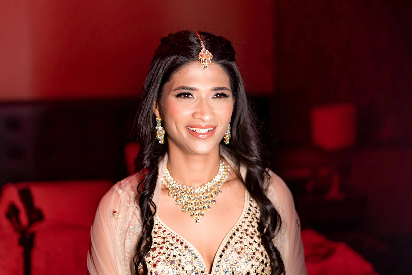 Medha smiles in front of a red sofa with a fine tikka and a gold necklace