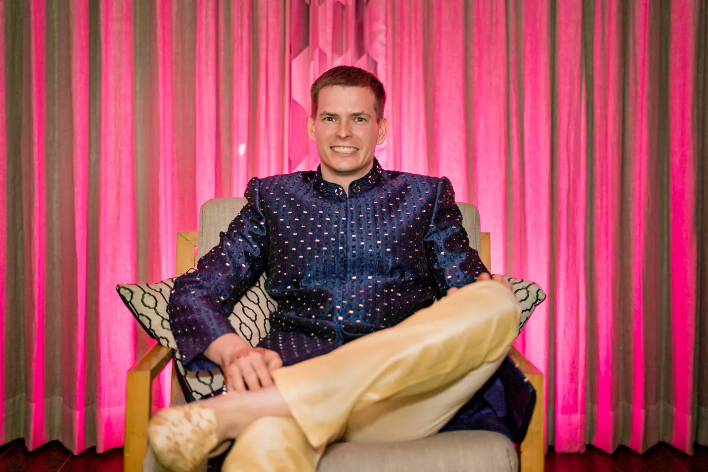 Stefan sits relaxed in an armchair against a bright pink curtain, one ankle over his knee