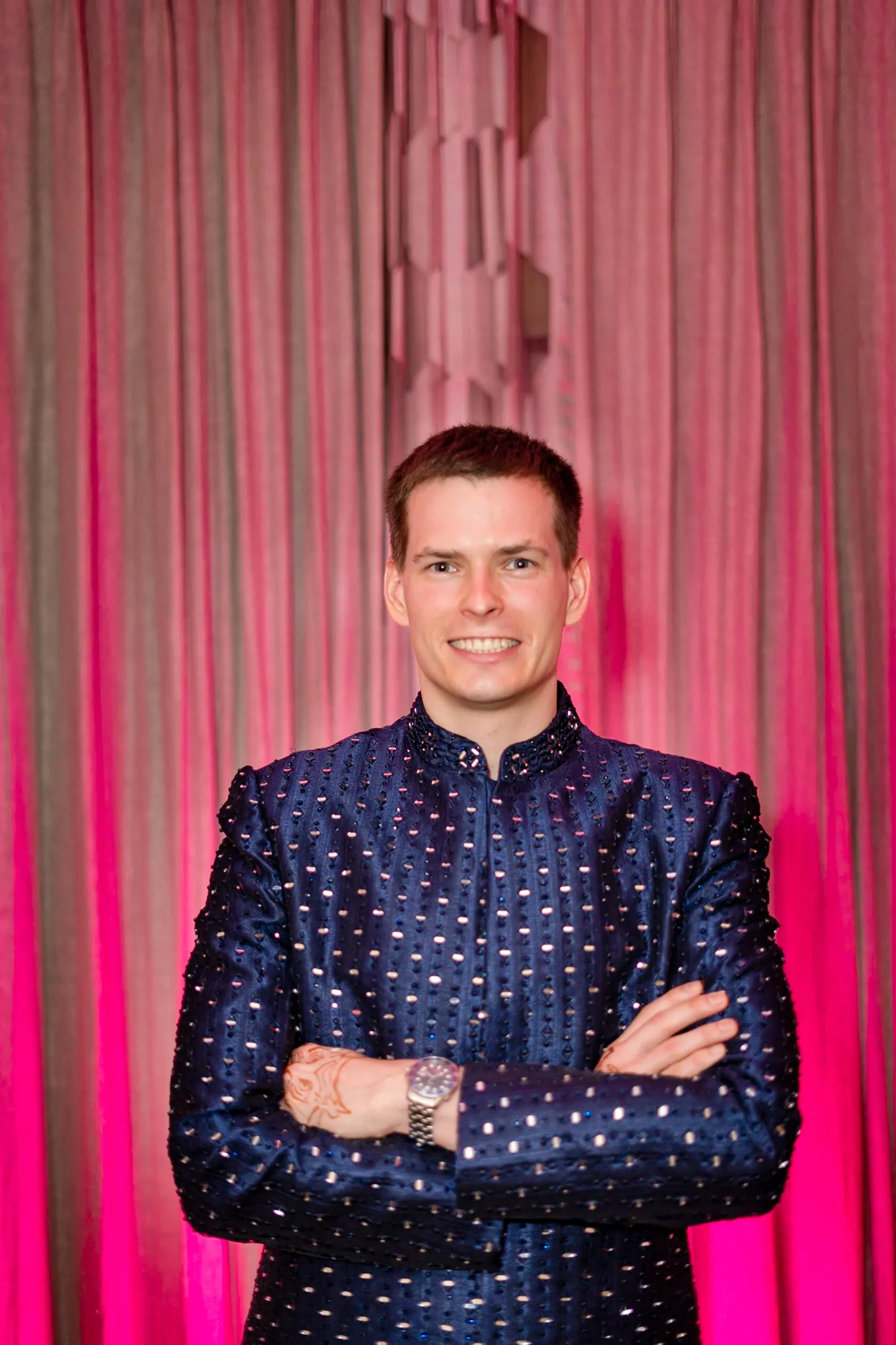 Stefan stands with arms folded and a wide grin against a pink curtain