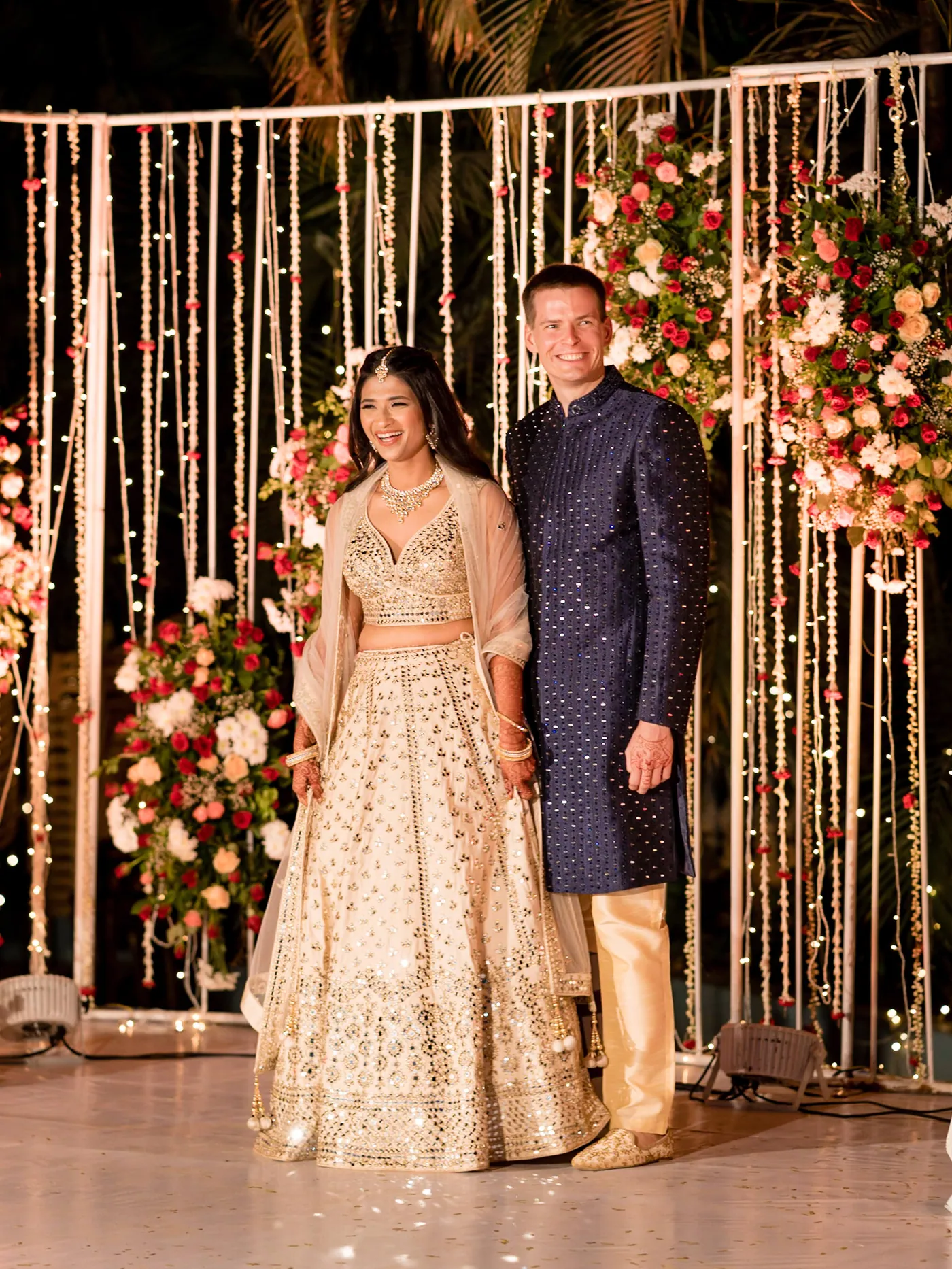 Medha in a cream lehenga and Stefan in a navy sherwani stand in front of a curtain of hanging lights