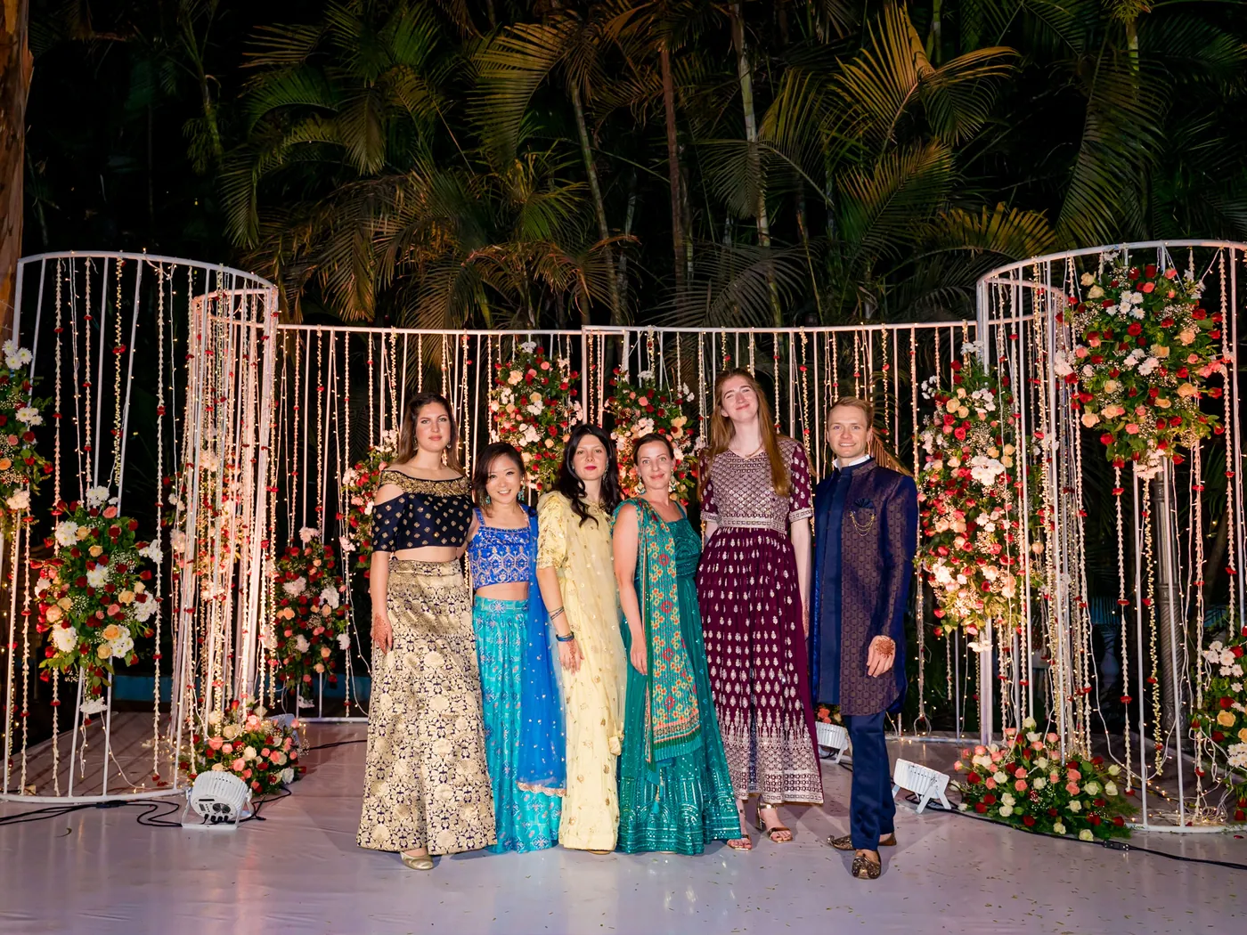 A row of friends in teal, gold and plum outfits pose in front of the glowing backdrop