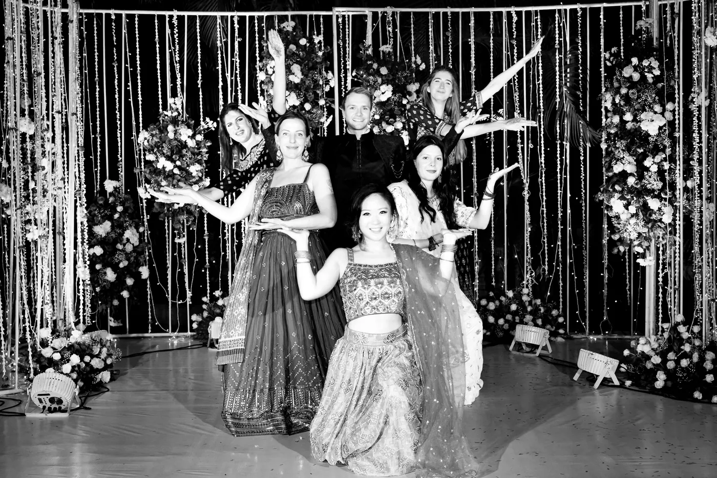 In black and white, friends throw their arms out in a playful pose in front of the strings of lights