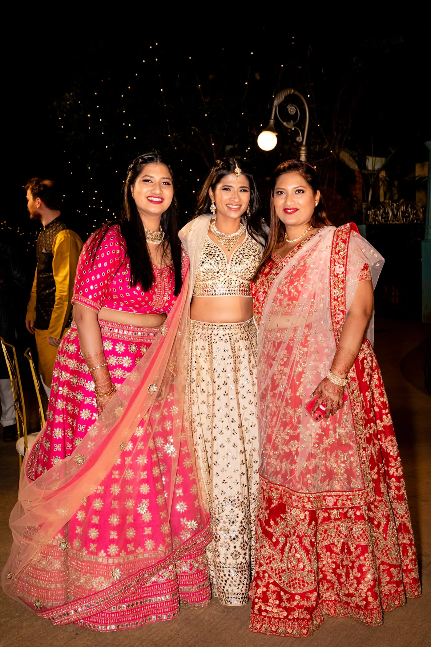 Medha stands between two of her sisters in pink and red lehengas, all three grinning