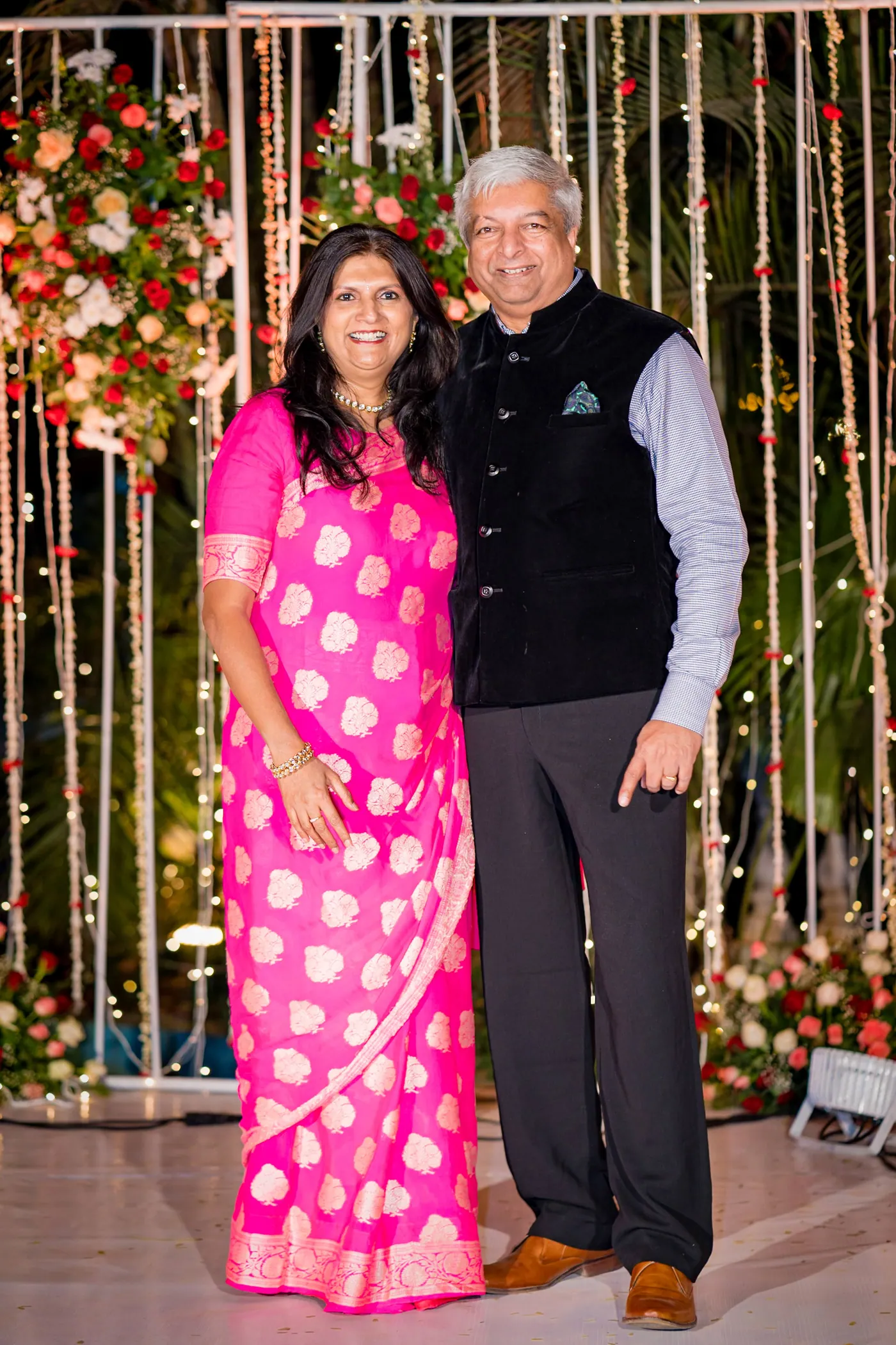 A woman in a bright pink sari and a man in a dark waistcoat pose in front of the backdrop