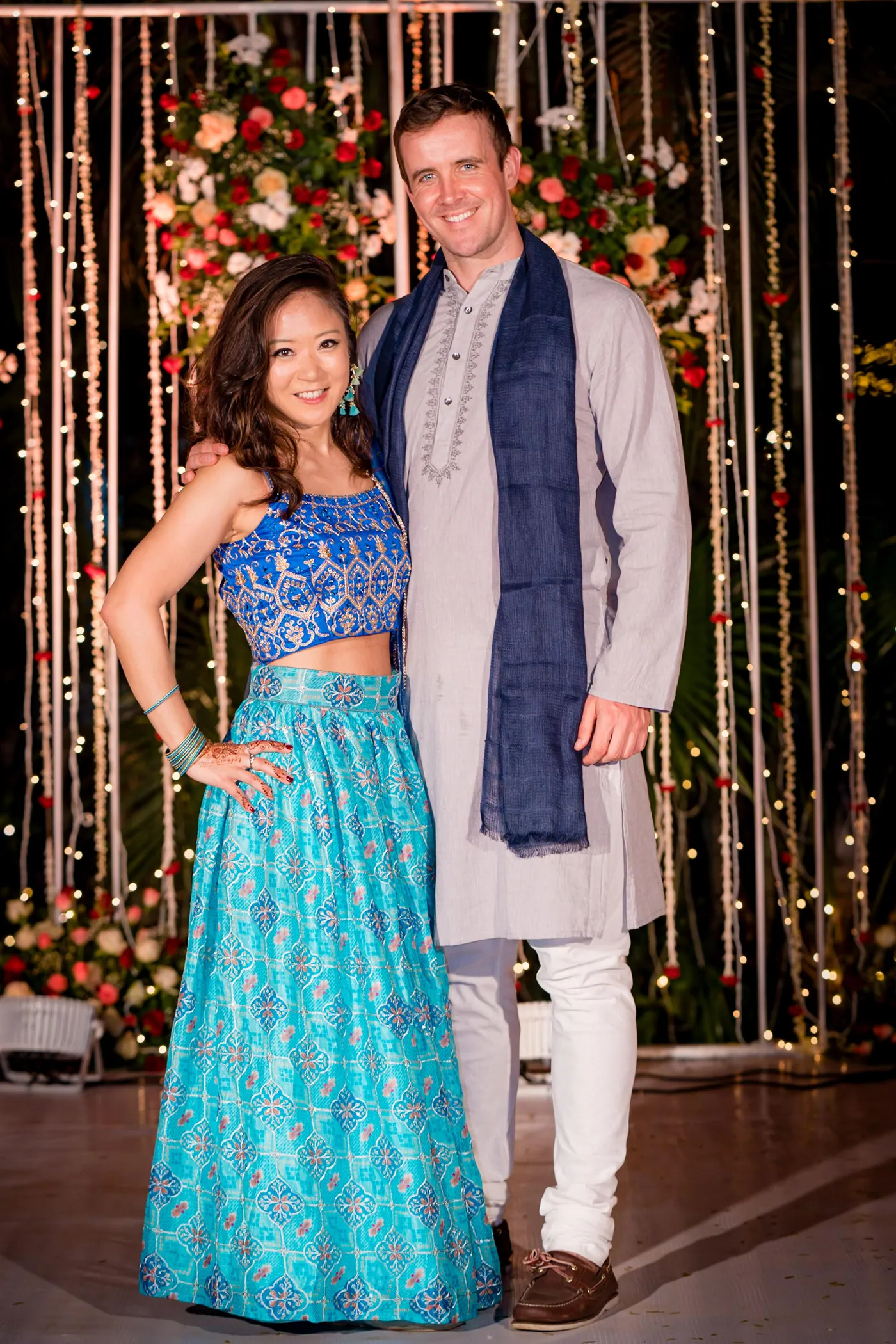 A woman in a turquoise lehenga and a man in a pale kurta and navy shawl pose arm in arm