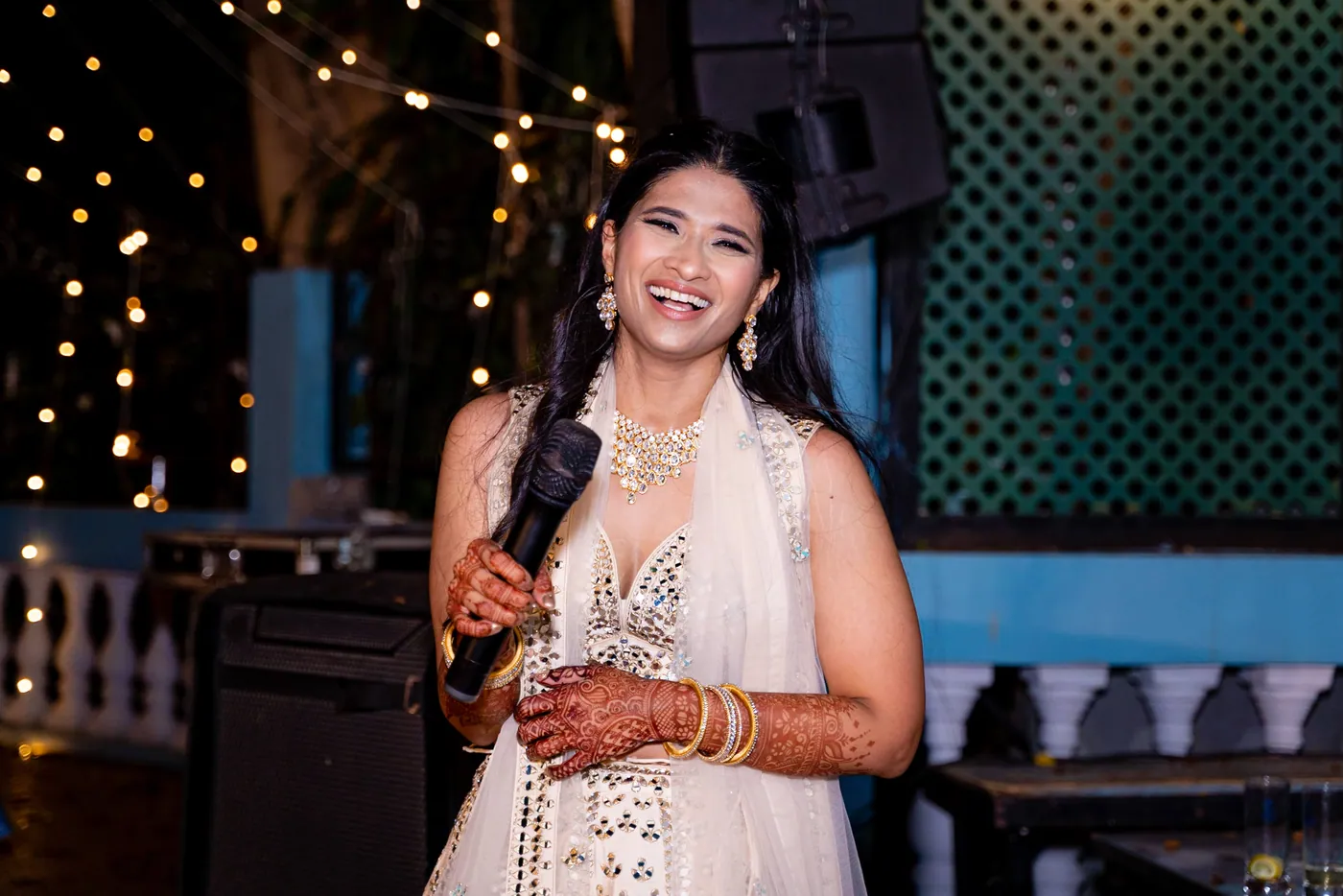 Medha smiles and laughs holding a microphone in a cream lehenga