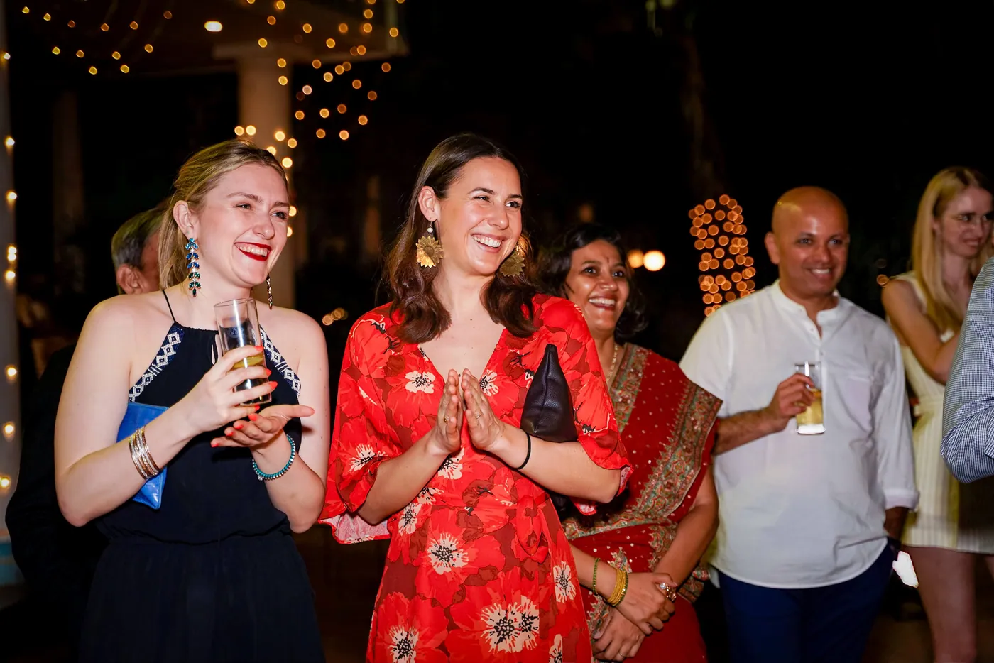 Three women, including one in a red floral top, clap and smile as they watch