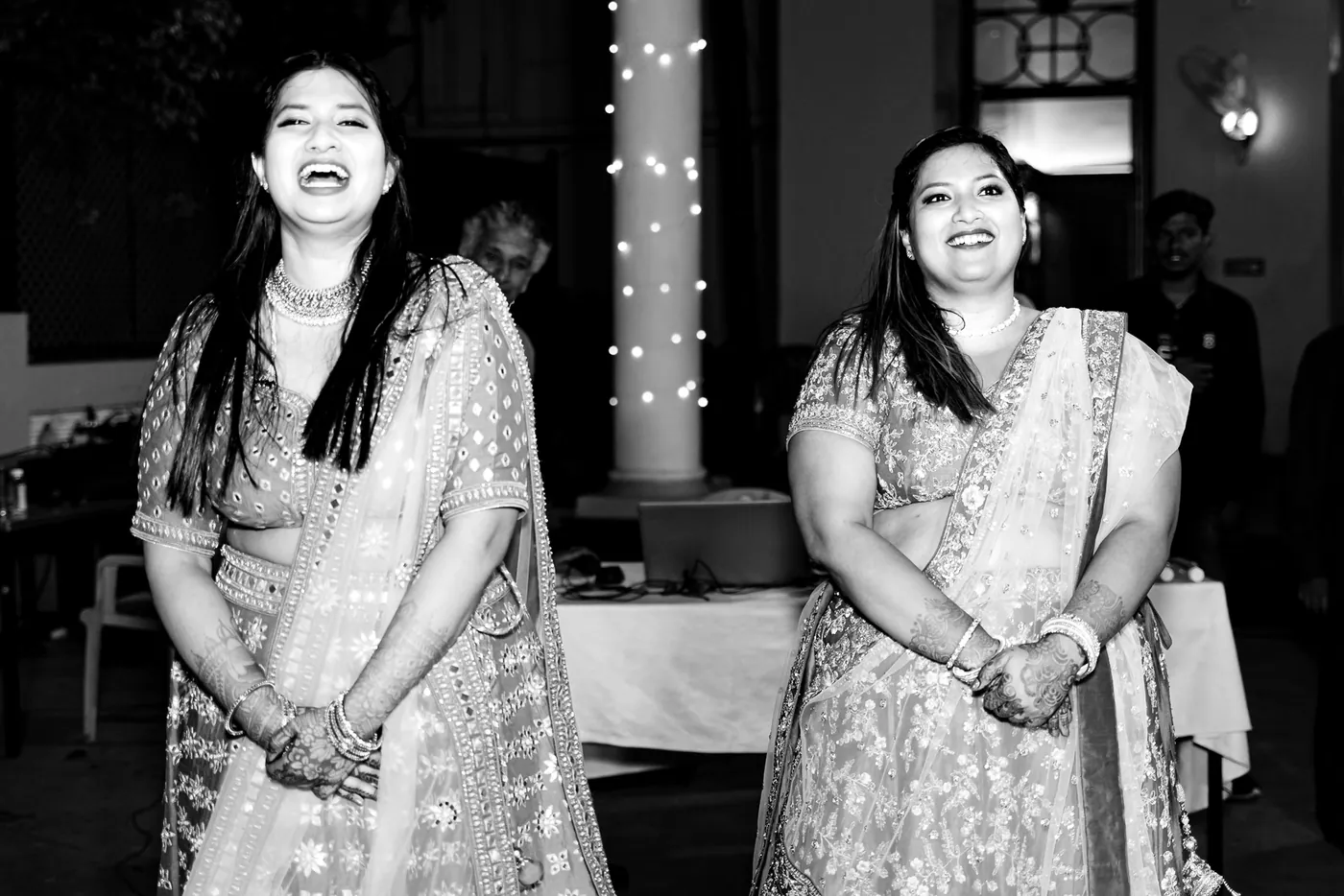 In black and white, two women in lehengas laugh out loud with their hands together