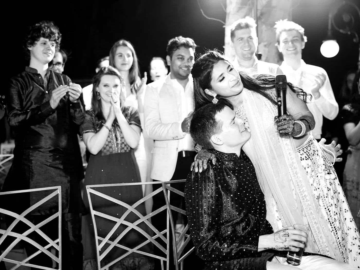 In black and white, Medha leans back with a microphone beside a kneeling man in a dark kurta