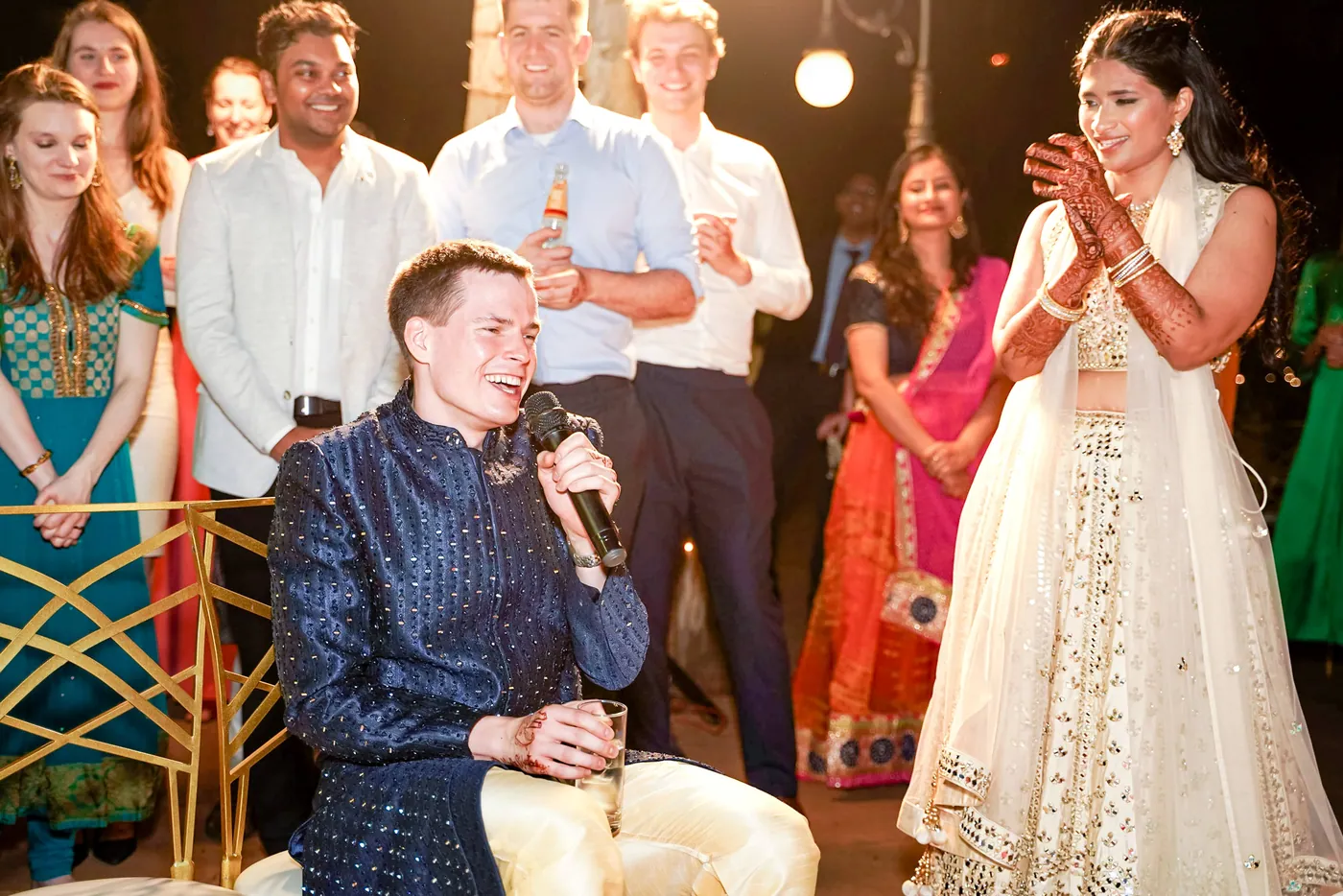 Stefan sits in a navy sherwani holding a microphone while Medha stands smiling beside him and friends look on