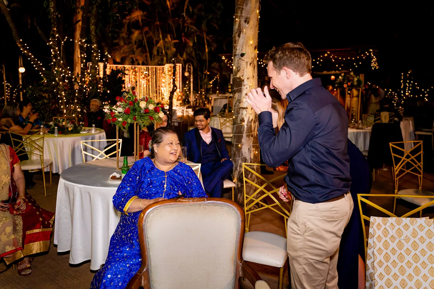 A woman in a bright blue kurta sits at a table while a man in a navy shirt claps nearby