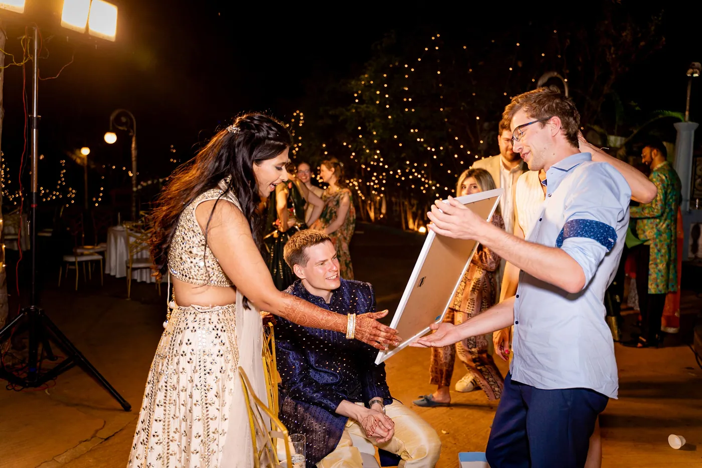 Medha holds a framed board as a man in a pale blue shirt and seated Stefan in navy laugh together