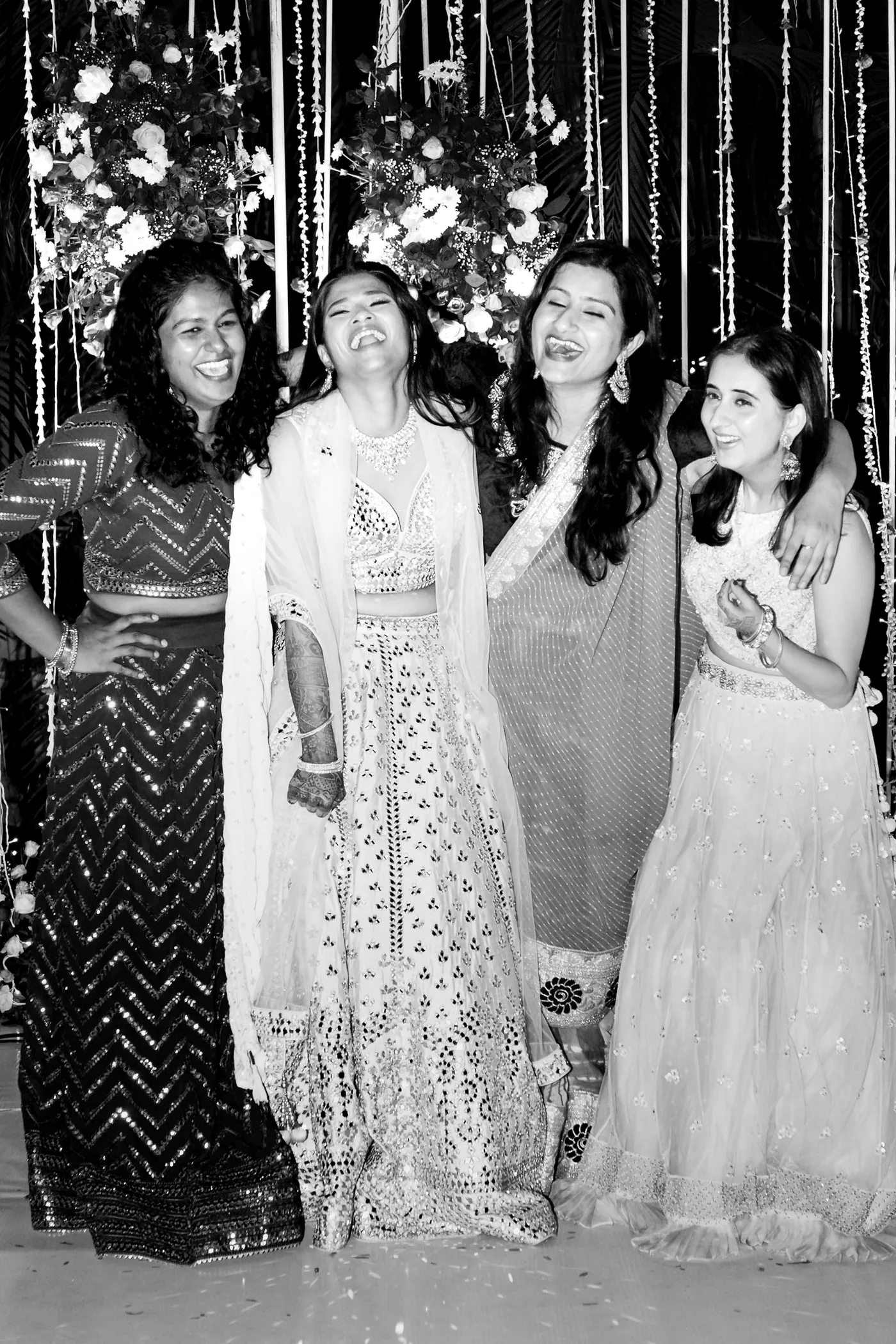 In black and white, four women in lehengas and a sparkling top laugh together with their arms around each other