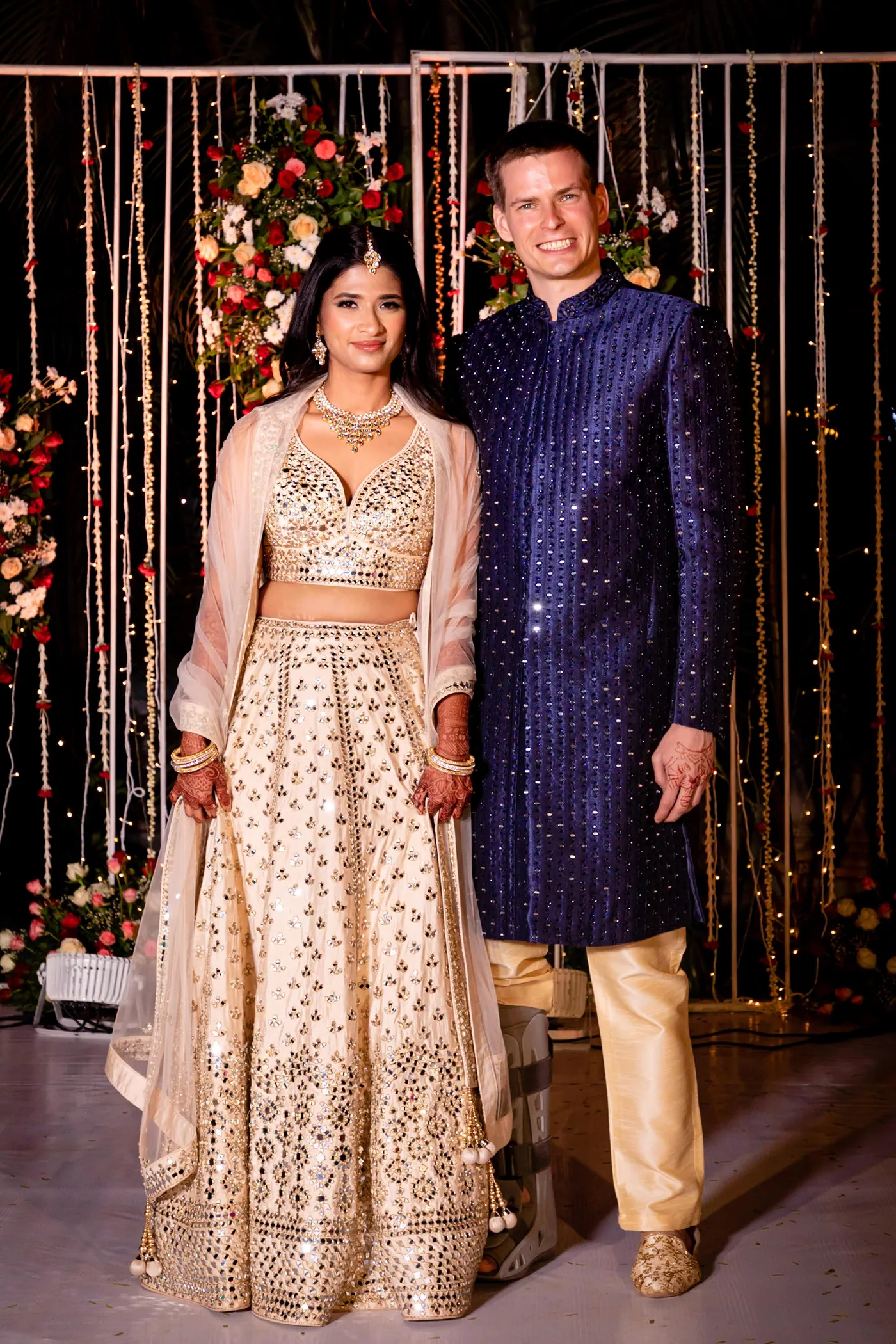 Medha and Stefan pose hand in hand on the stage, flower arrangements and lights behind them