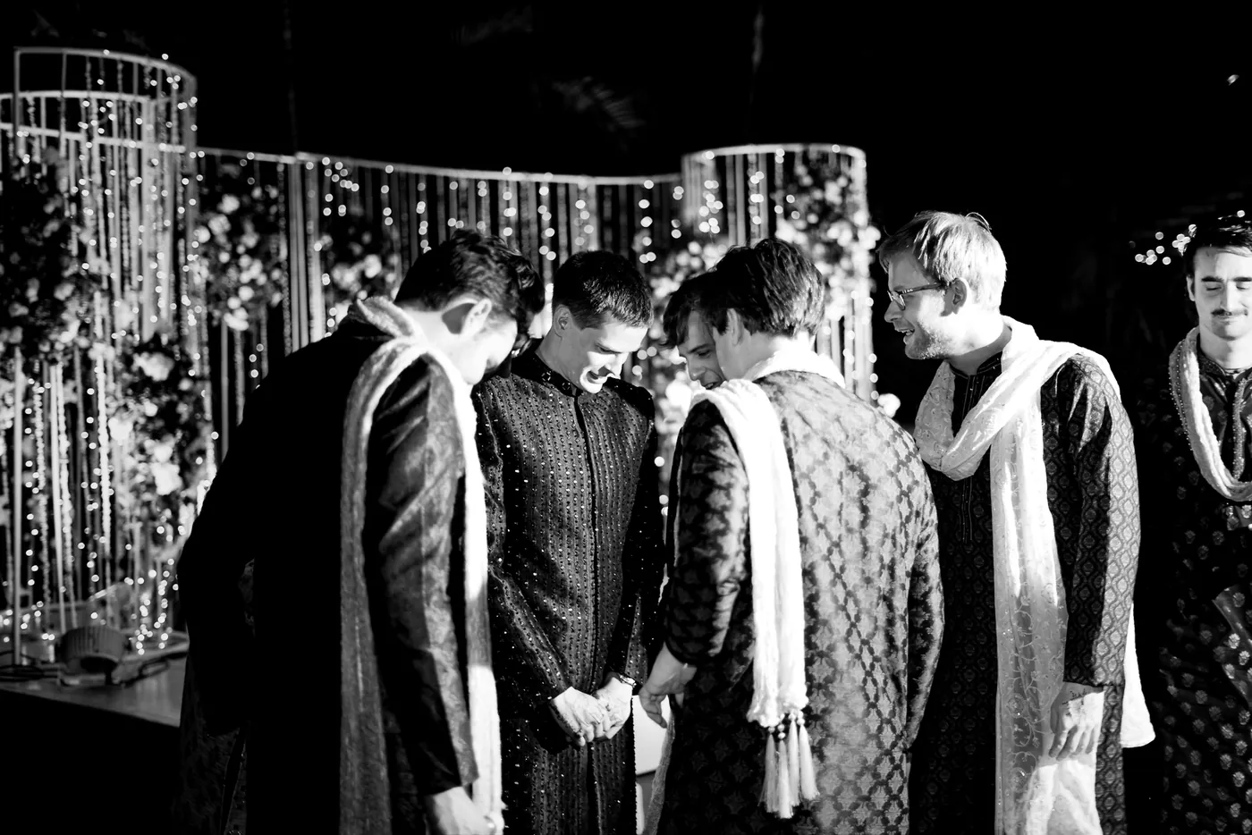 In black and white, a huddle of men in kurtas lean in together under strings of lights