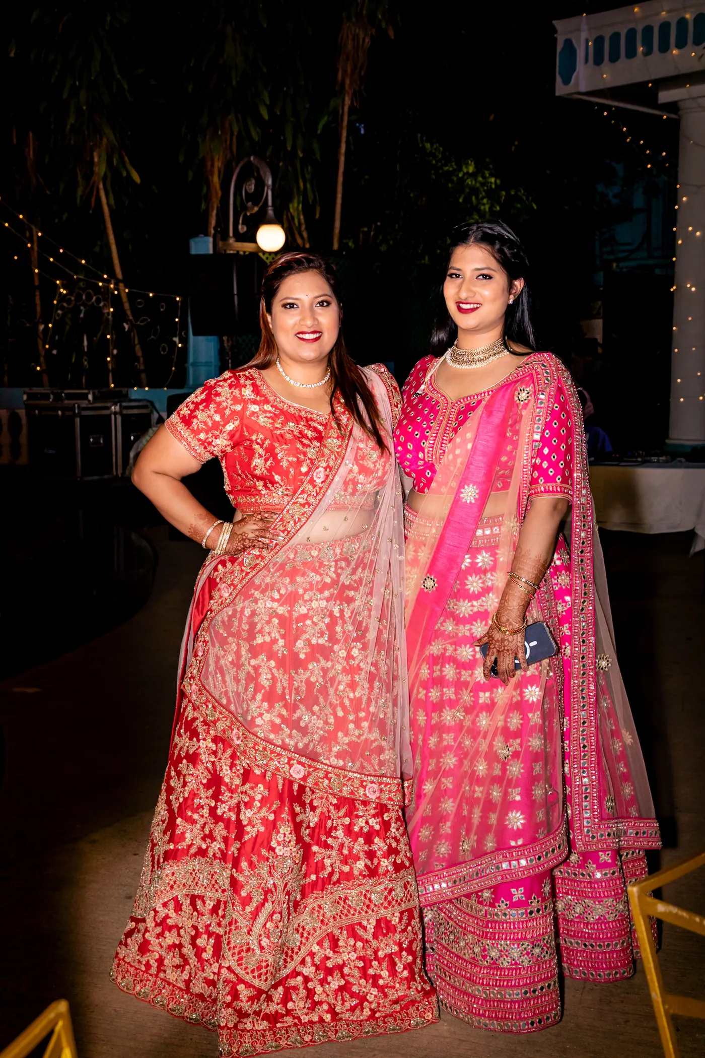 Two women in red and pink lehengas pose for the camera at night with lights behind