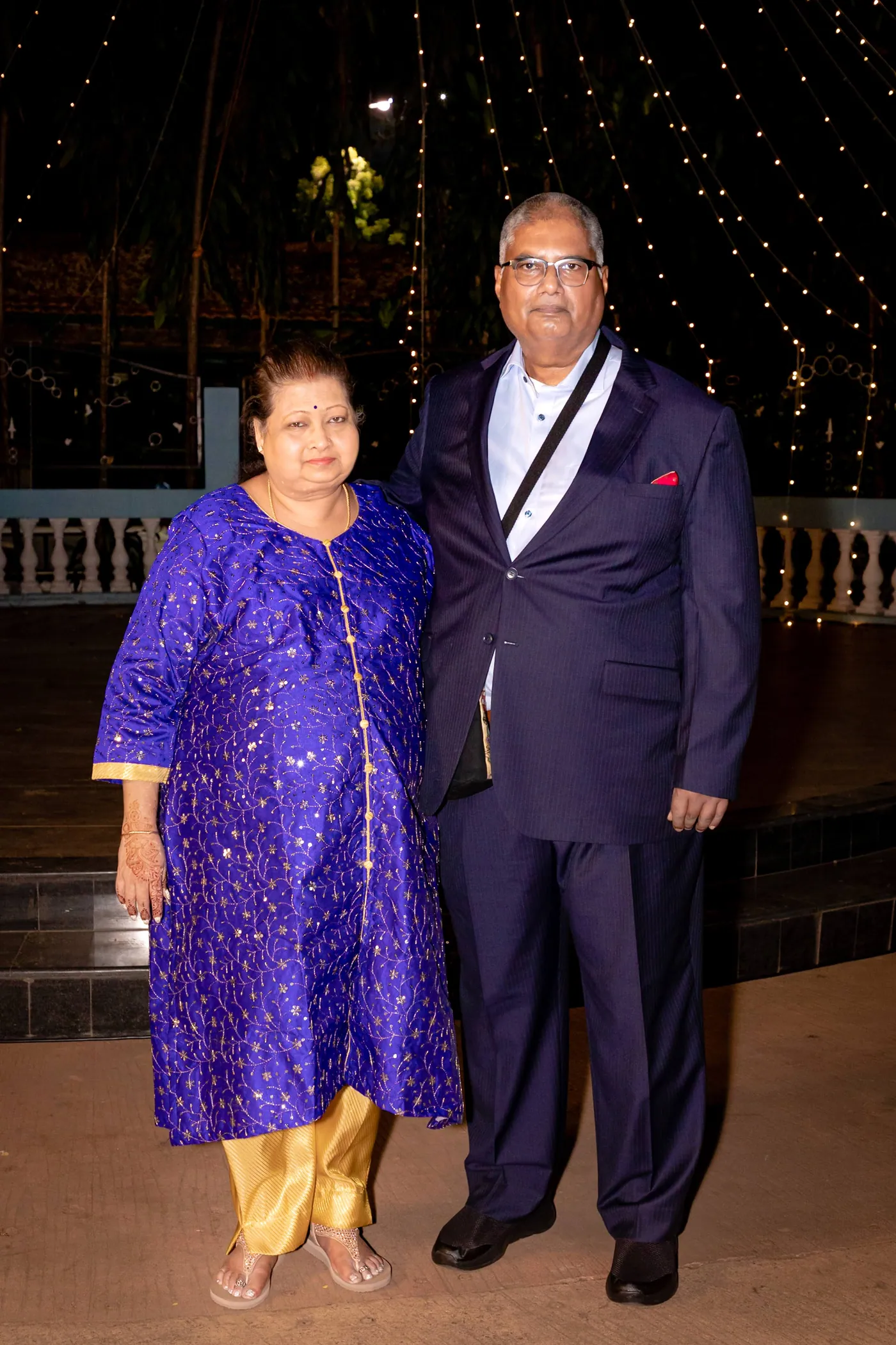 A man in a dark suit stands next to a woman in a violet kurta with the club lit behind