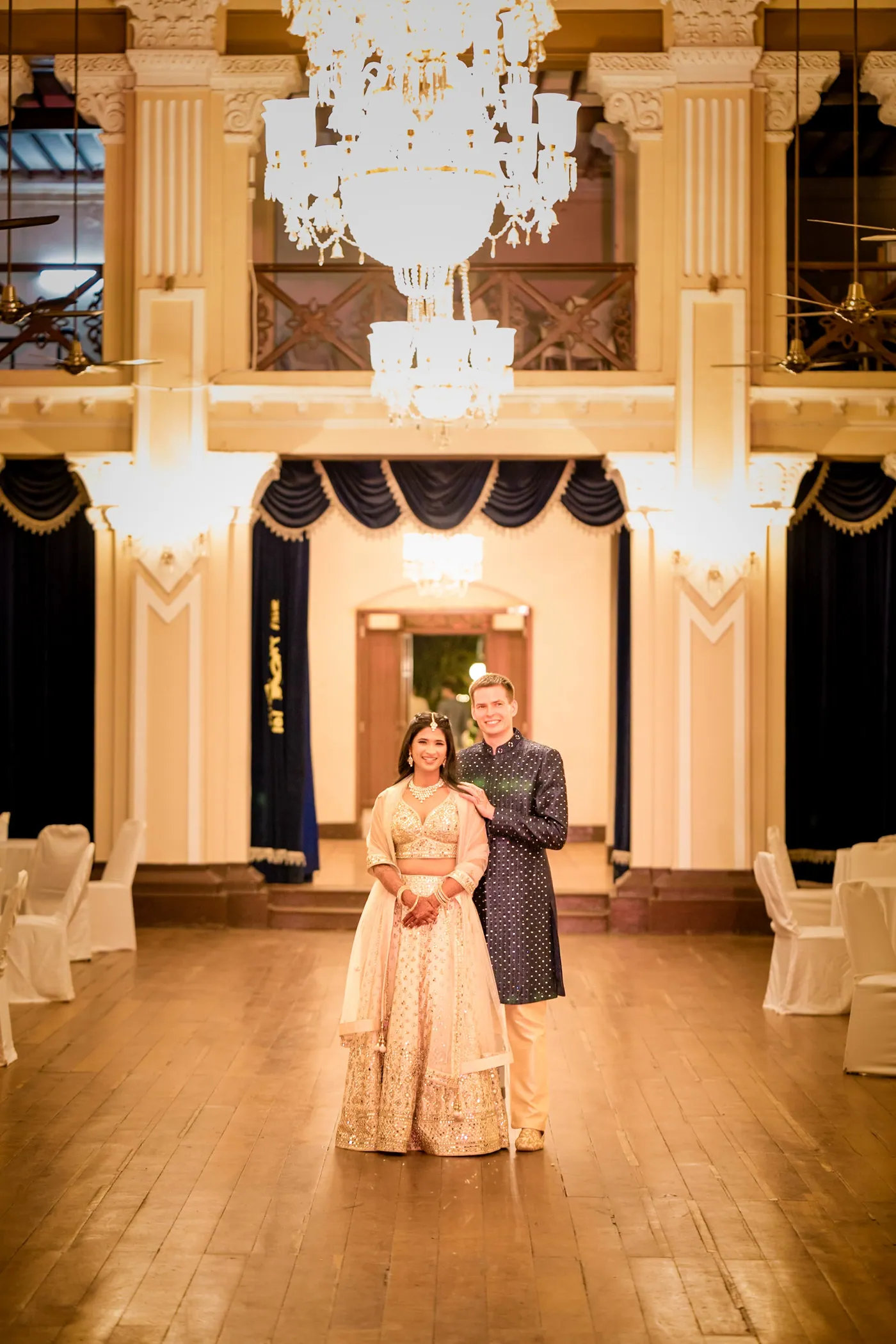 Medha and Stefan walk down the long colonial club hall toward the camera under glowing chandeliers