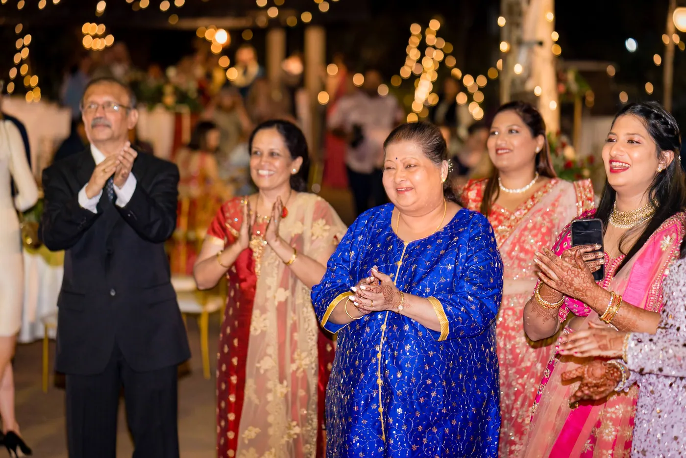 Guests clap and smile beside a woman in a bright blue kurta at the start of the evening