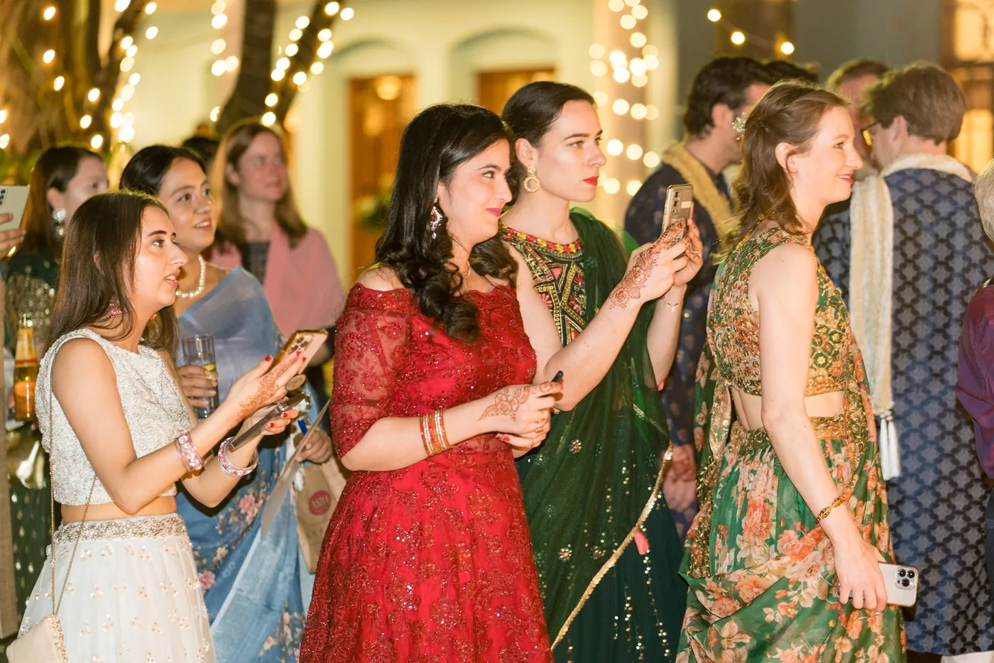 Women in lehengas and saris crowd together, one holding up a phone, under strings of lights
