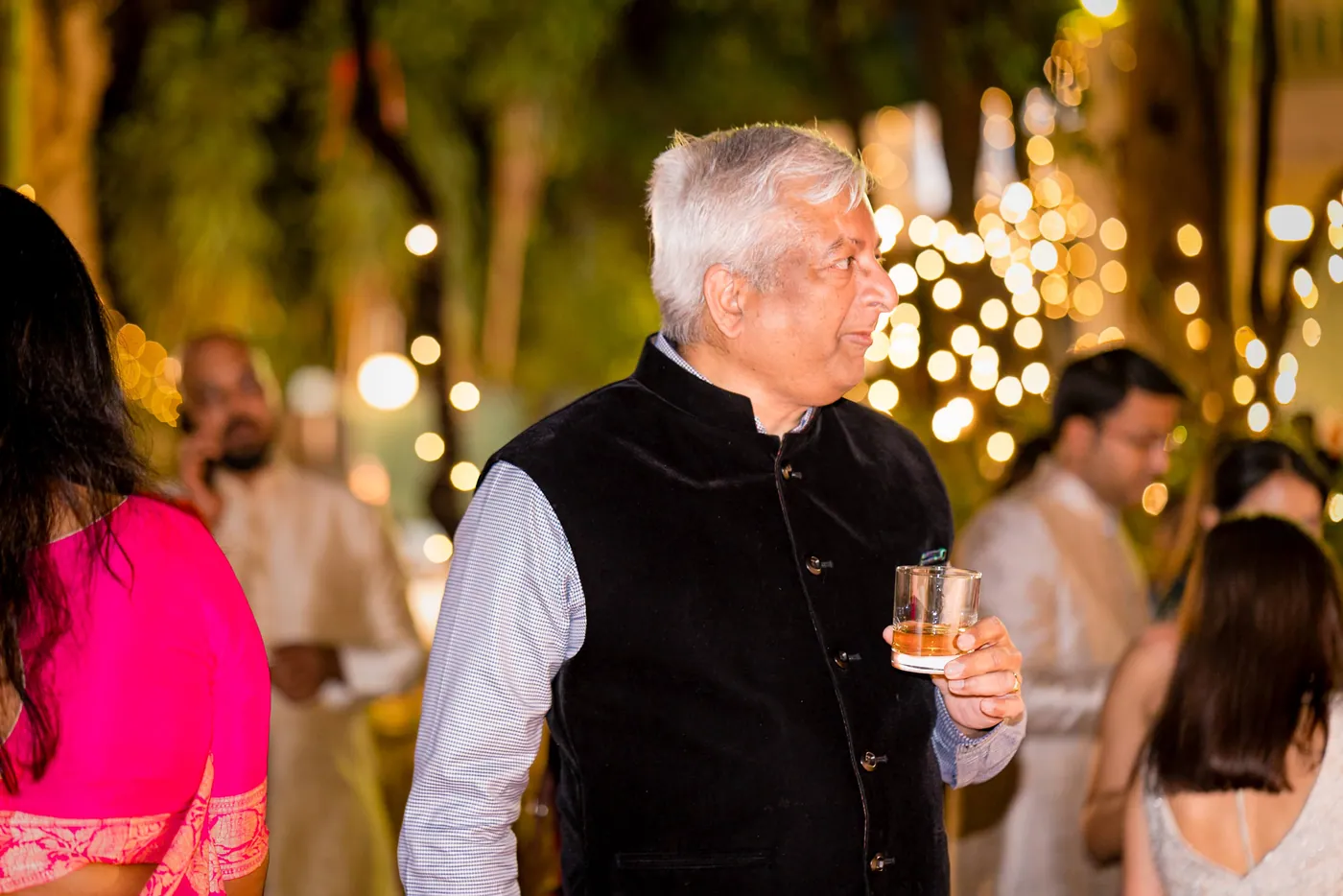 An older man in a black waistcoat holds a glass and talks as fairy lights bokeh behind him