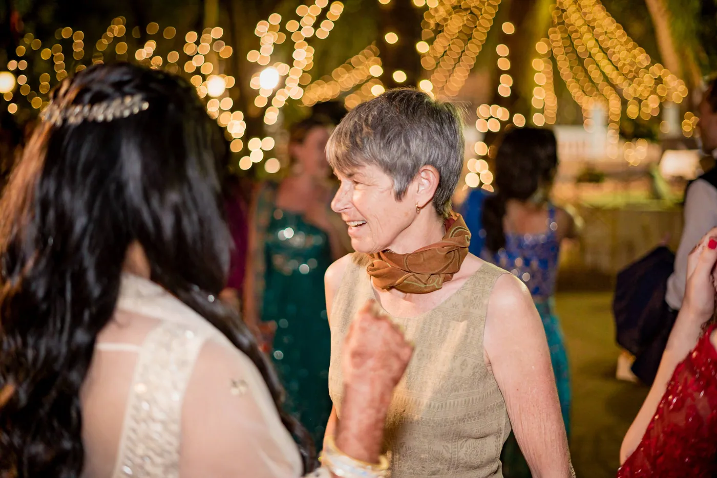 Medha leans in to talk with a grey haired woman in an orange scarf while fairy lights glitter behind
