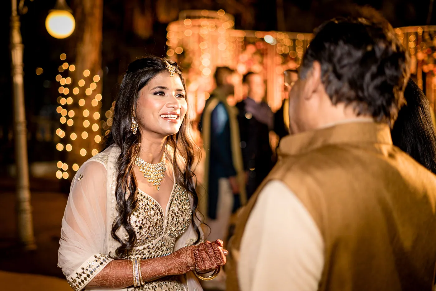 Medha in a cream lehenga with henna on her hands smiles past a man in a gold jacket