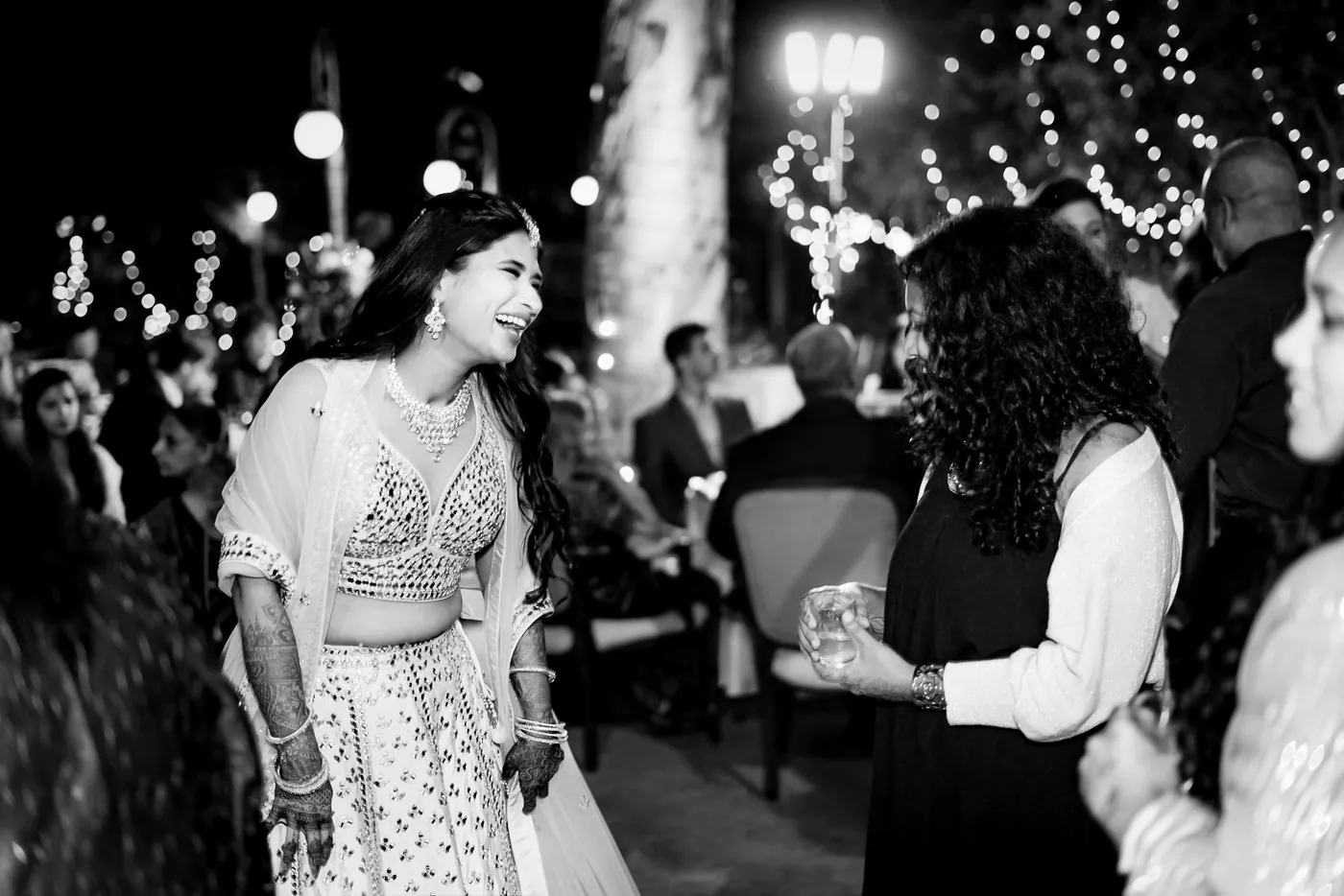 In black and white, Medha laughs and turns to greet a woman as the party fills up behind her