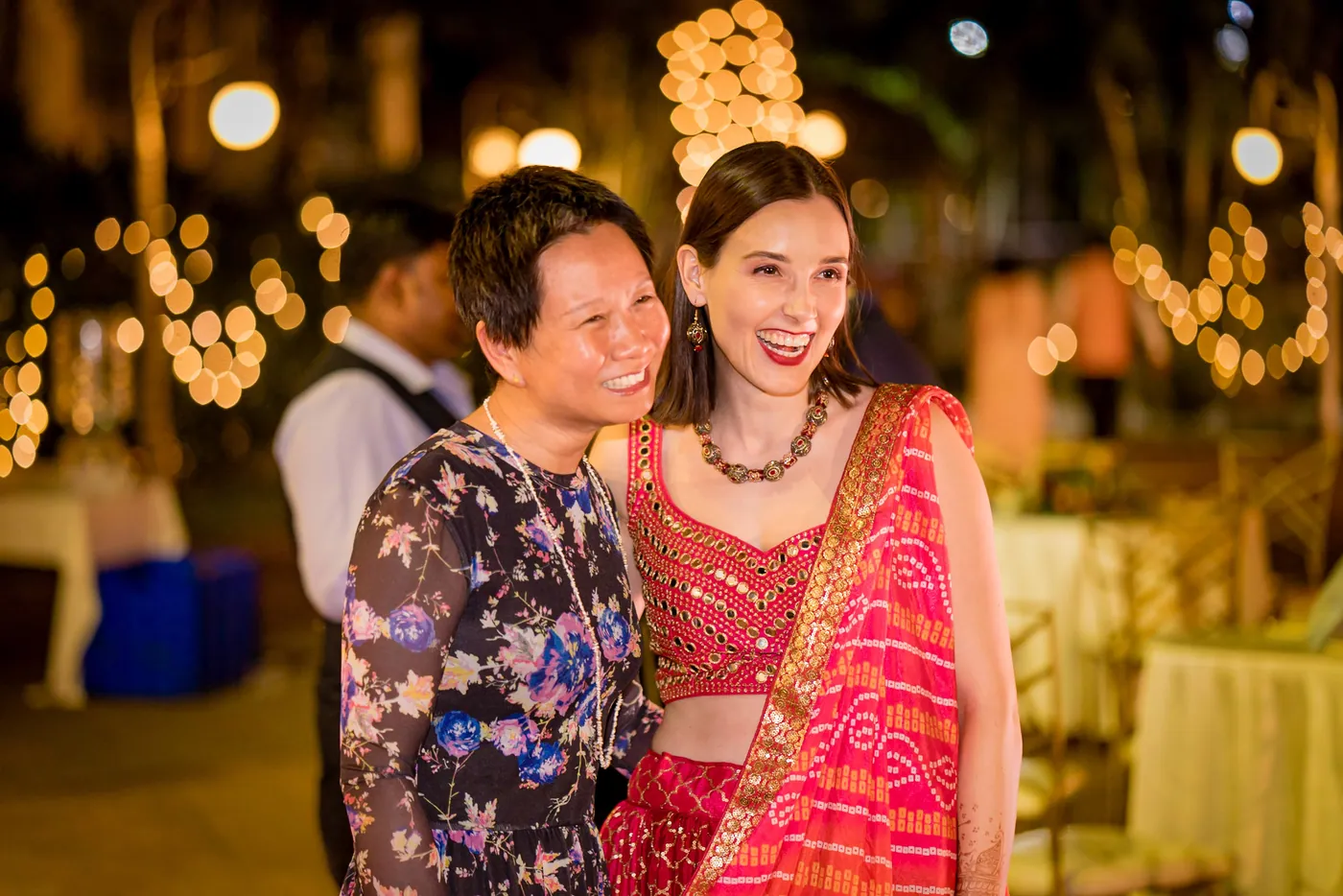 A woman in a patterned jacket and a woman in a red crop top smile together close to the lens