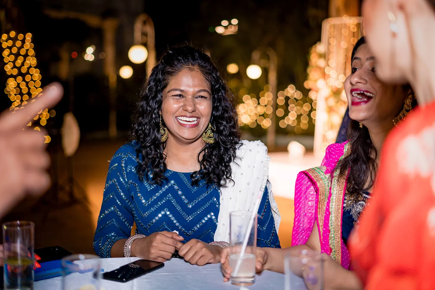 A woman in a blue top smiles broadly at a table with a drink near her hand